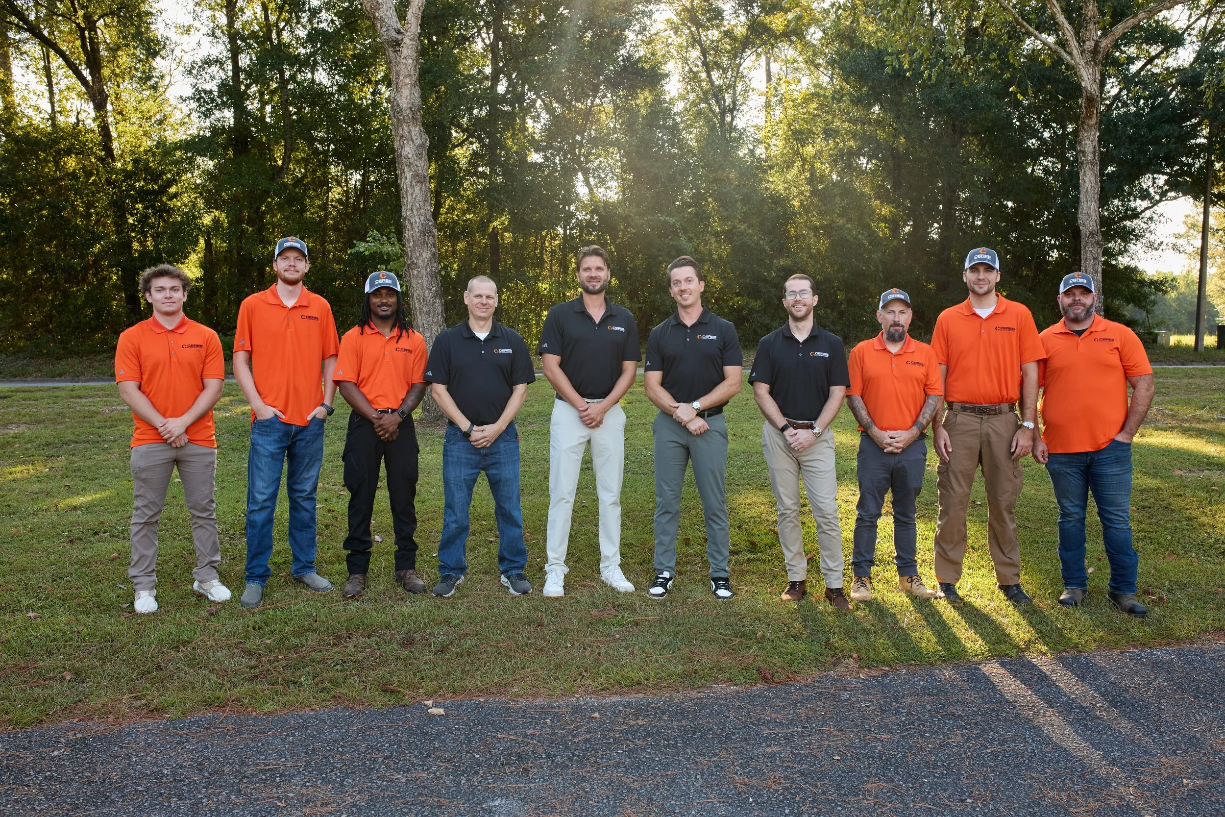 Group of ten people standing outdoors on grass in front of trees during sunset, wearing black and orange shirts with a company logo.