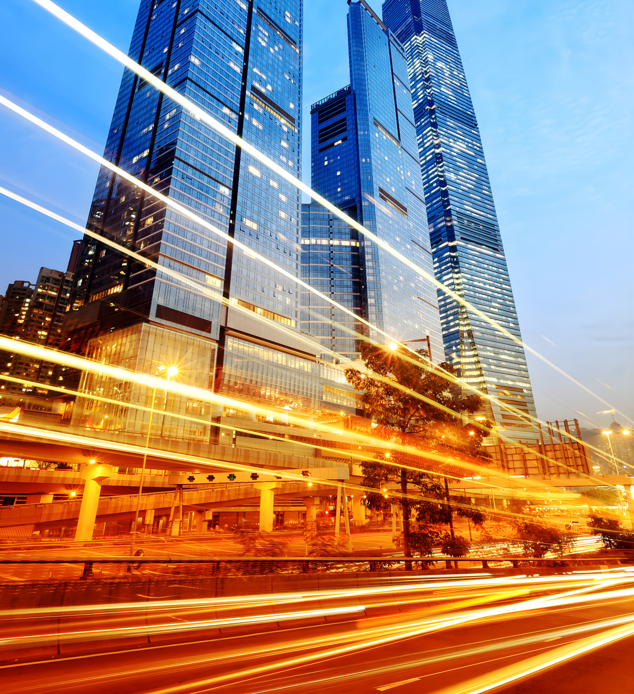 Nighttime cityscape featuring tall modern glass skyscrapers with light trails from moving vehicles on the street in front.