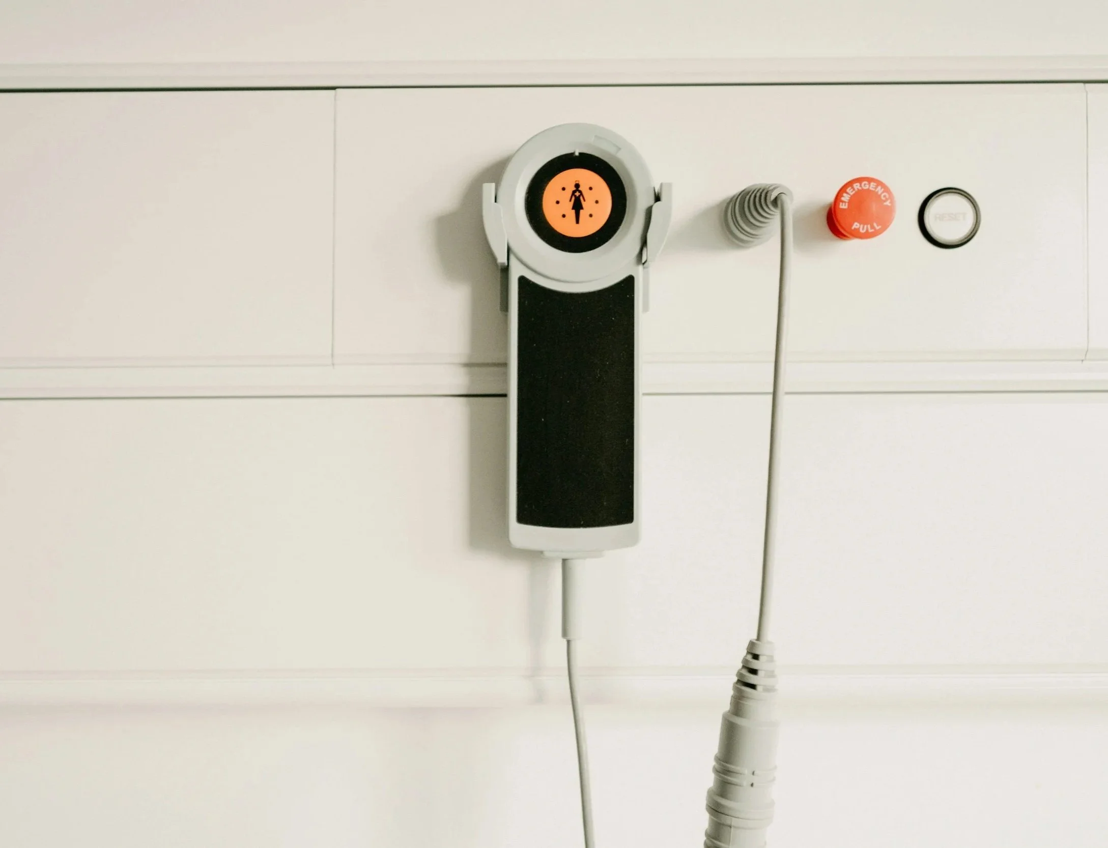 Medical equipment attached to a hospital wall, including a patient monitor with a symbol of a person, an emergency pull button, and a reset button.