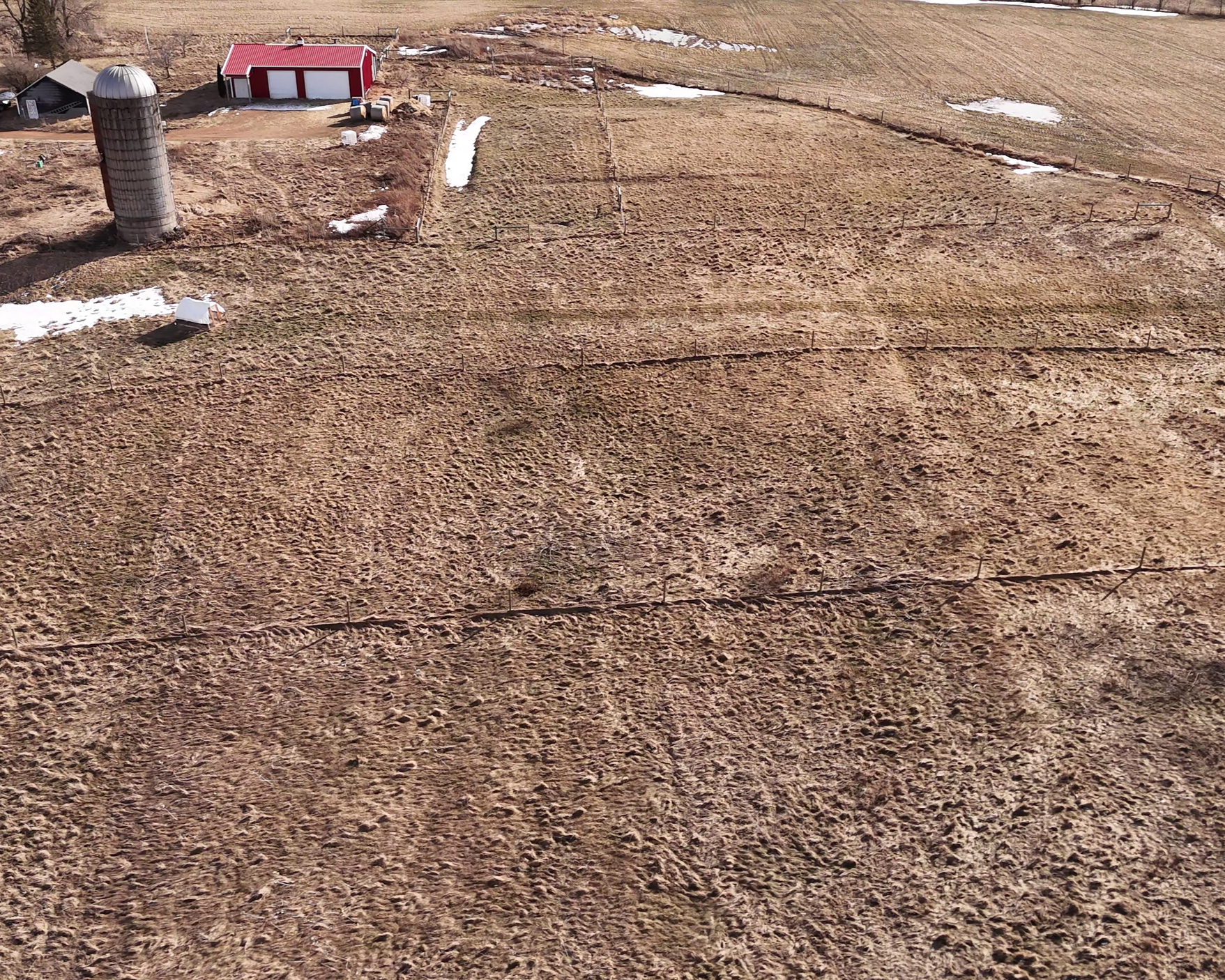 Aerial view of farmland with brown fields, patches of snow, a small barn, a silo, and fencing.