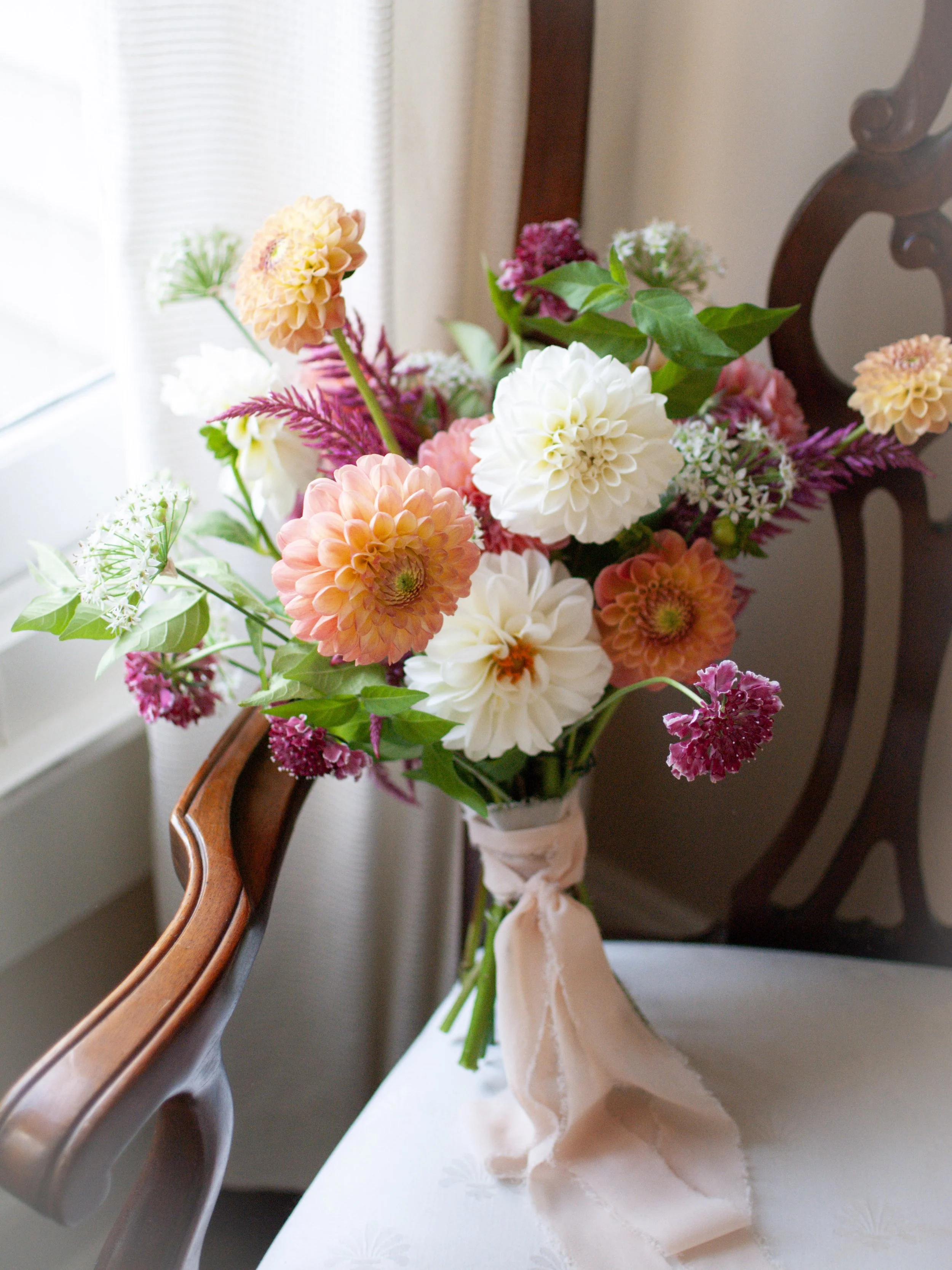 A bouquet of pink, white, and peach flowers, tied with a light pink ribbon, placed on a white table near a window with curtains. Designed by St. Louis Florist, Suncrest Blooms.