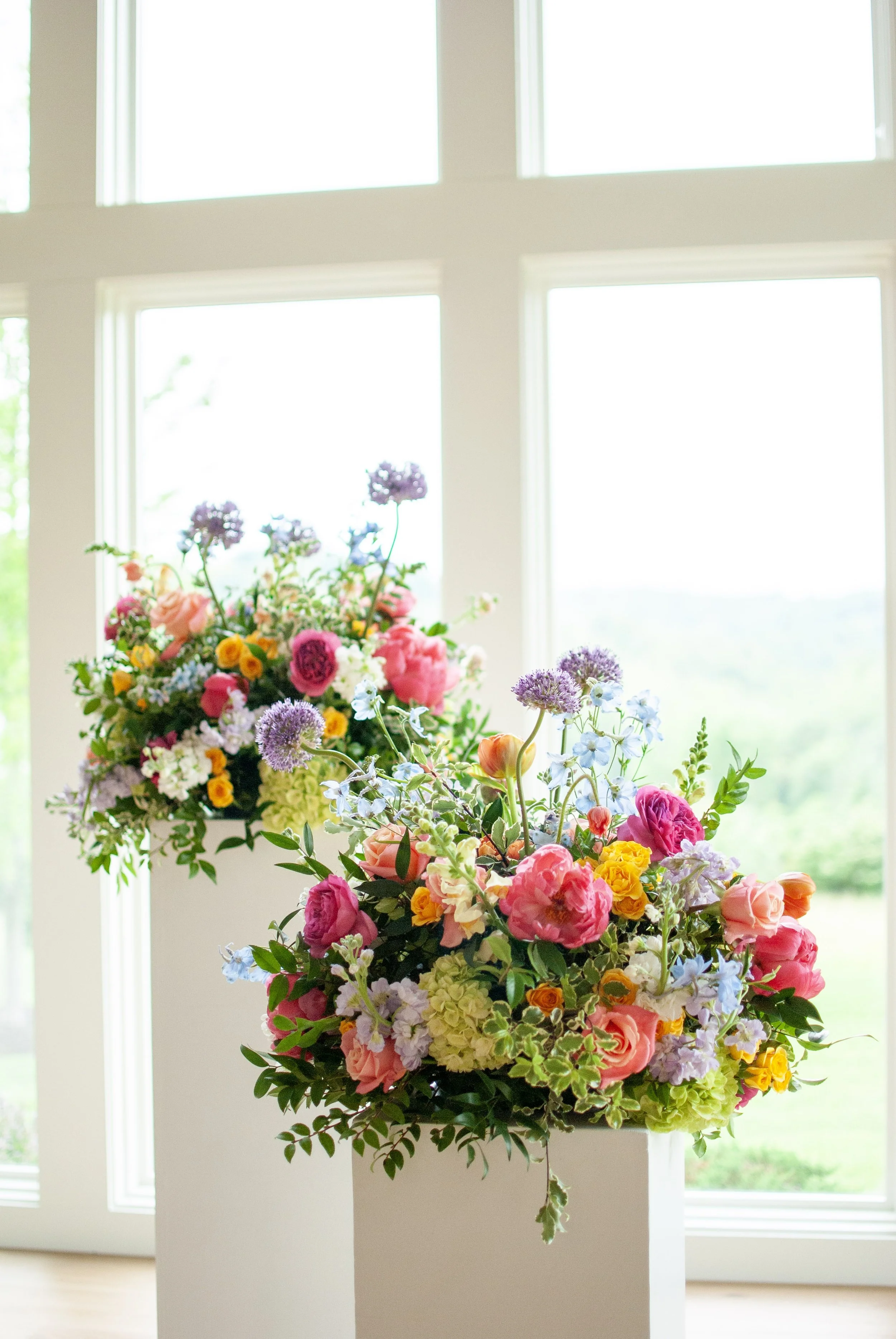 Two floral arrangements with colorful flowers including pink roses, purple alliums, yellow blooms, and green foliage, placed in front of large windows. Designed by St. Louis Wedding Florist, Suncrest Blooms.