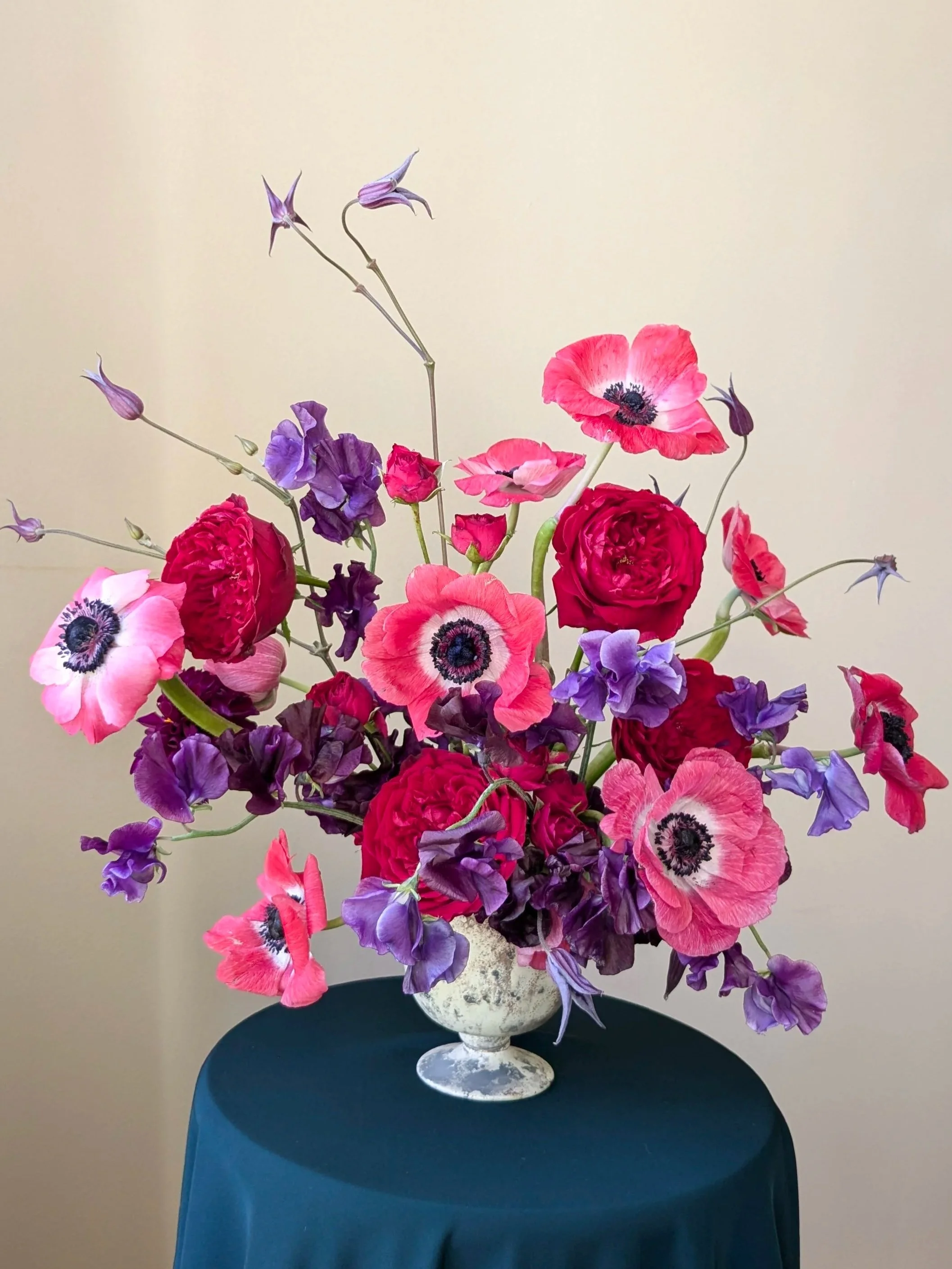 A bouquet of pink, purple, and red flowers in a white ceramic vase on a teal table against a plain beige wall.