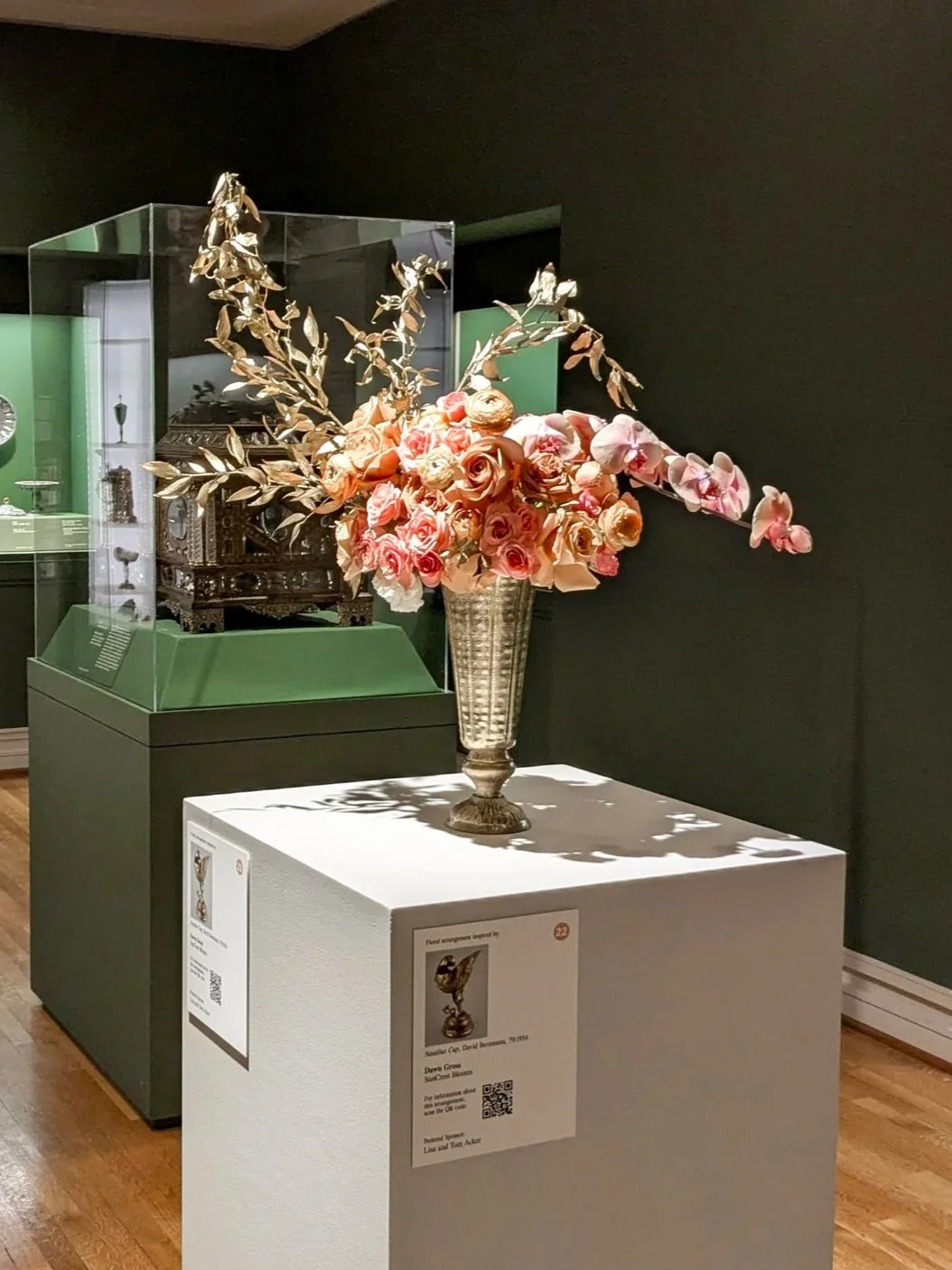 An ornate floral arrangement with pink roses, orchids, and gold-colored leaves in a silver vase, displayed on a white pedestal at a museum exhibit.