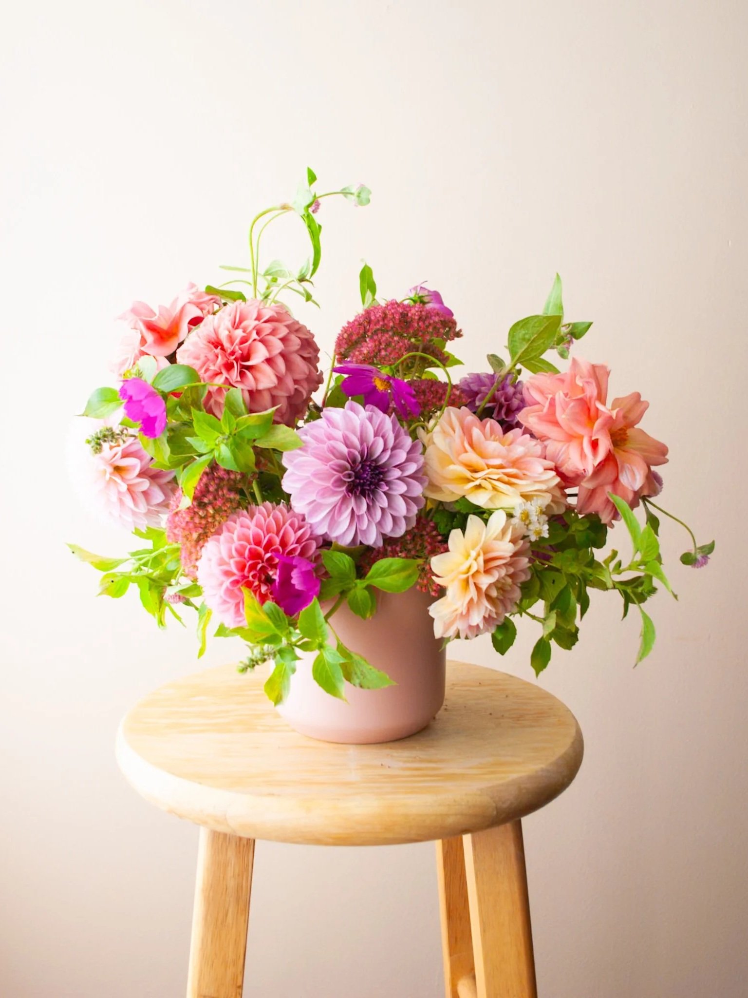 A pink vase filled with a colorful bouquet of pink, purple, and peach flowers, placed on a round wooden stool against a plain light-colored background.