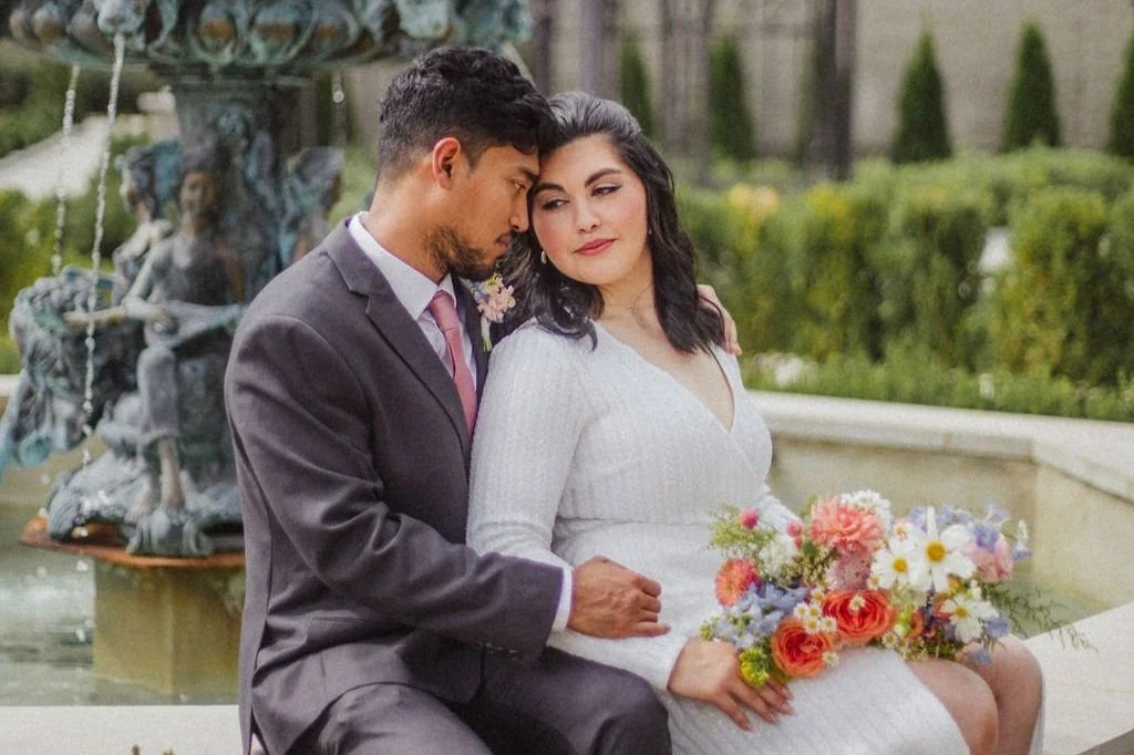Couple dressed in wedding attire sitting by a fountain, the woman holding a bouquet of flowers.