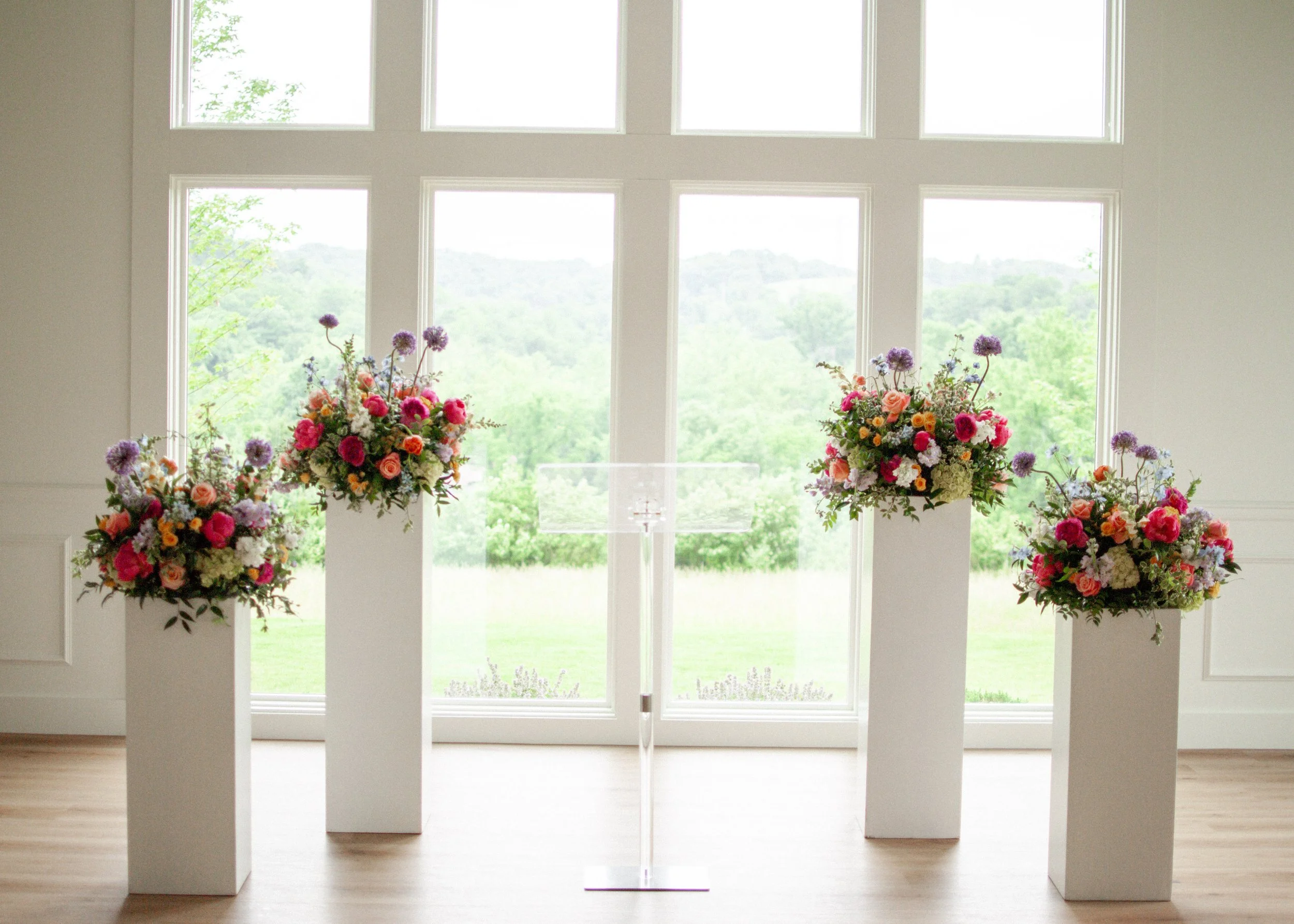 Bright room with large windows overlooking green landscape, decorated with three large floral arrangements on white pedestals. Designed by St. Louis wedding florist, Suncrest Blooms. Venue: Westwind Hills.