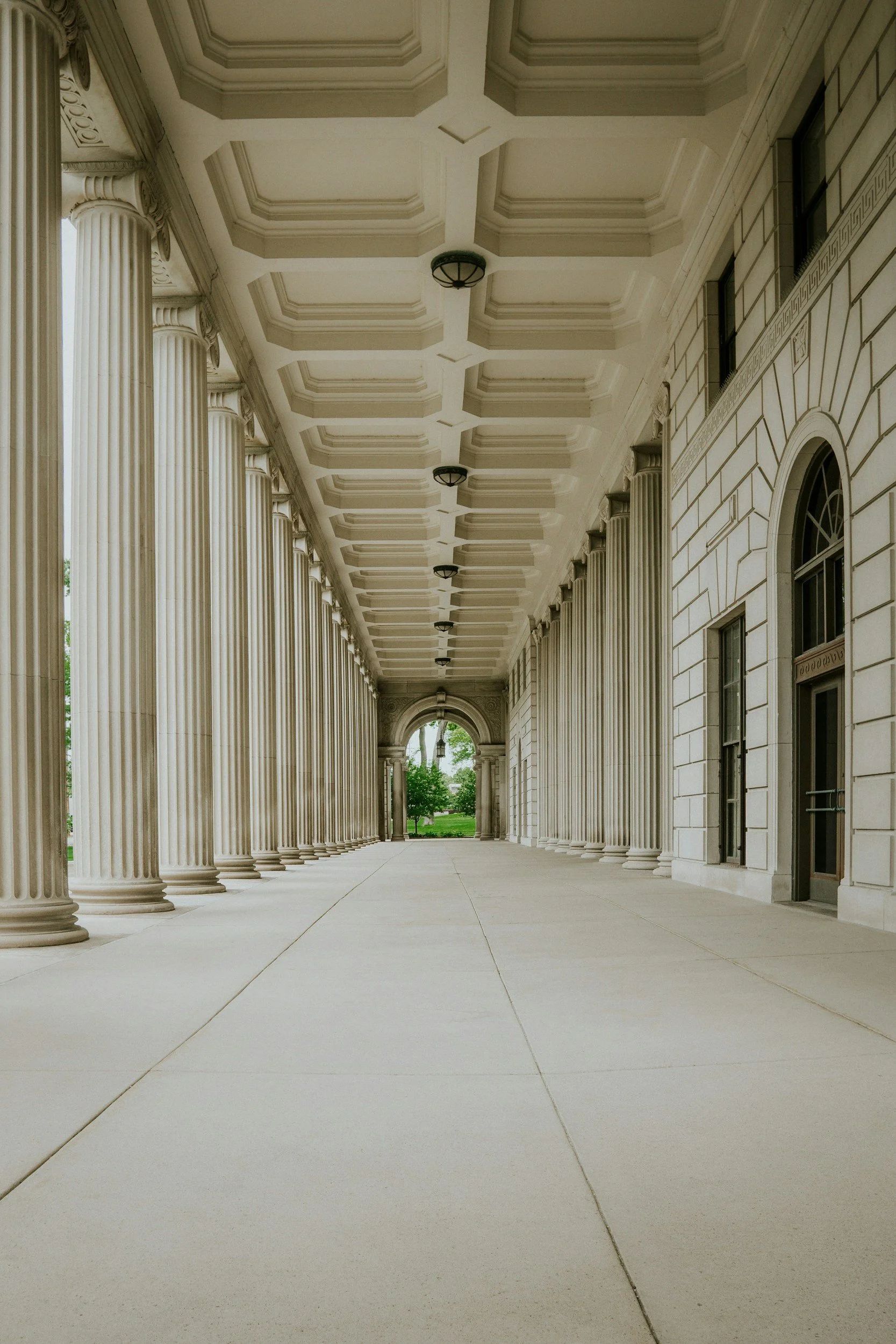 A covered walkway with tall, white classical columns on the left and a white building with arched windows on the right. The ceiling has a coffered design with evenly spaced, dark light fixtures. At the end of the walkway, there are green trees and a lamppost visible through an archway.