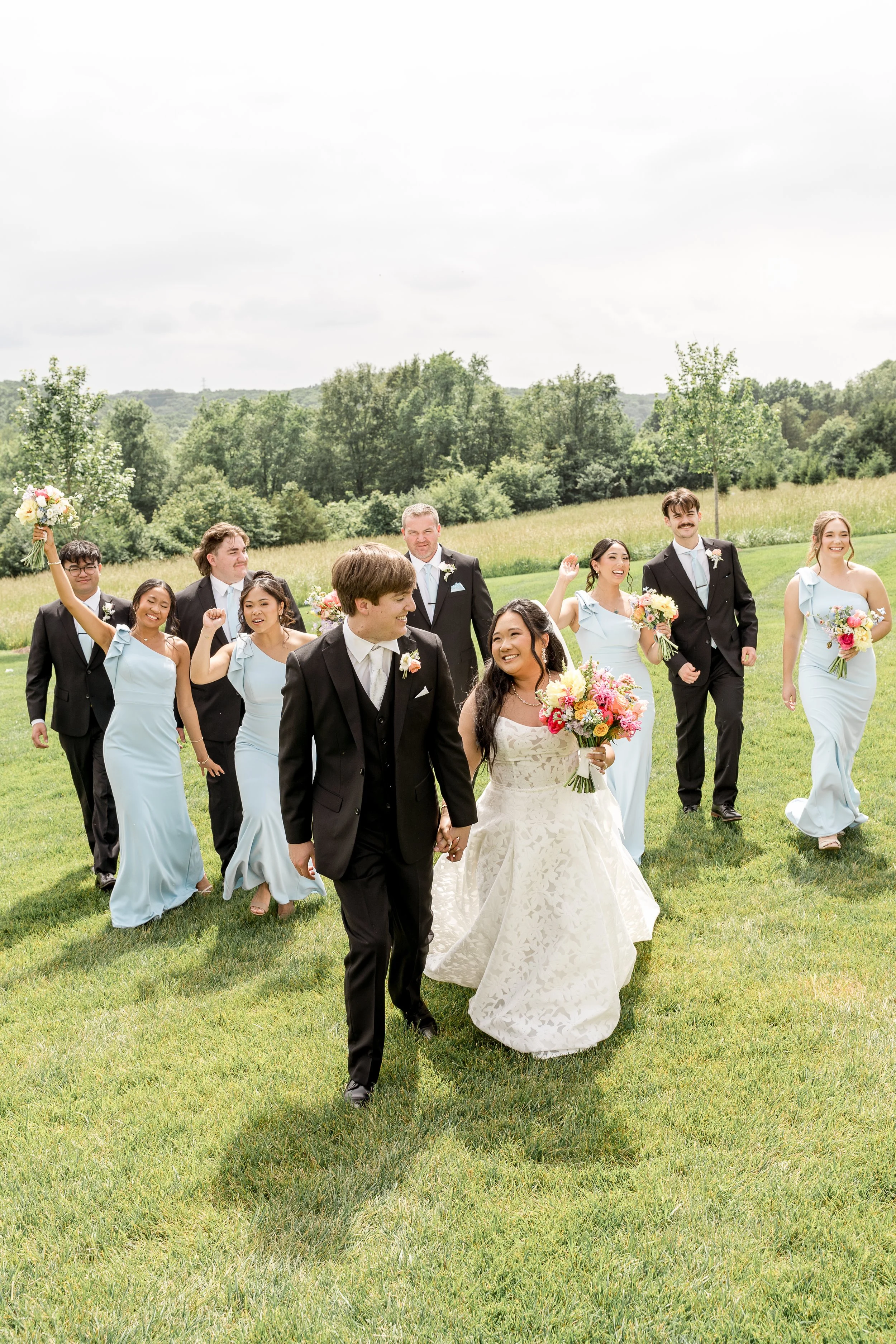 Bridal party walks on a green field smiling and cheering for the bride and groom. Bride is holding a vibrant bouquet designed by St. Louis florist, Suncrest Blooms.