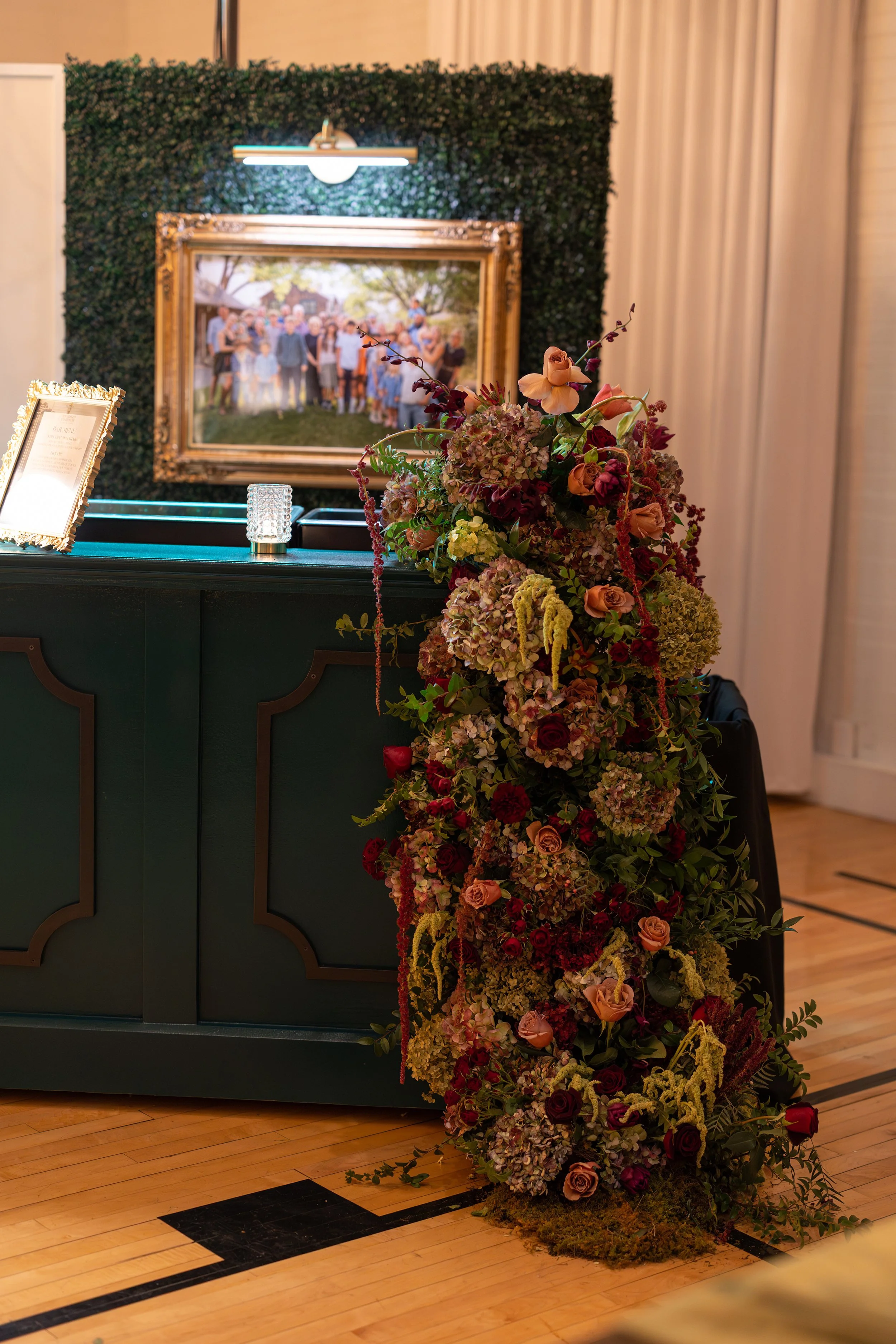 A floral bar cascade with pink, red, and purple flowers flowing down from the top of a bar to the ground. A framed group photo is on the wall behind, decorated with a green hedge. Designed by St. Louis Event Florist, Suncrest Blooms.