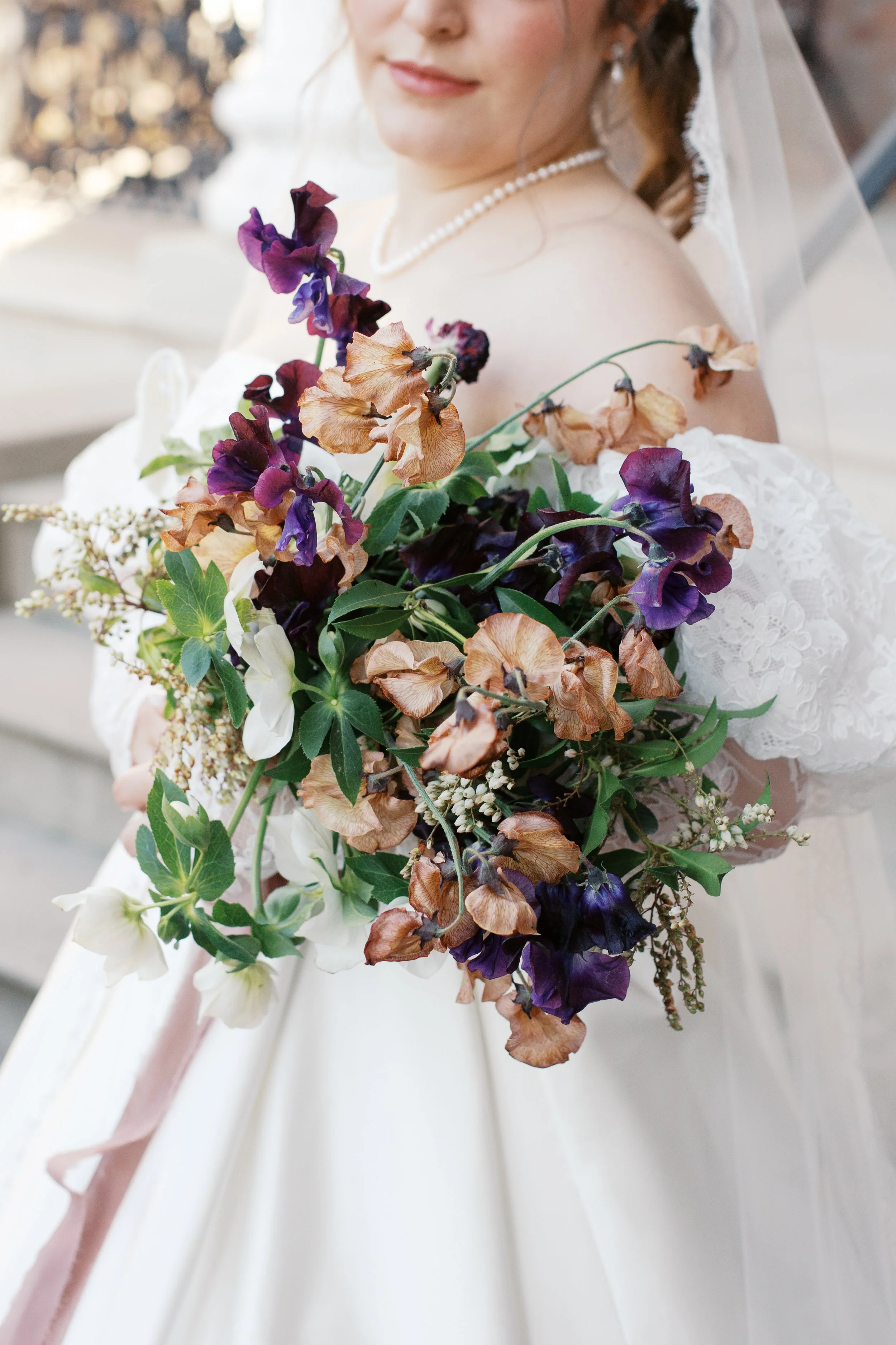 A bride holding a bouquet of dried and fresh flowers, including purple and beige blooms, with a bride in a white dress and veil visible in the background. Designed by St. Louis wedding and event florist, Suncrest Blooms.