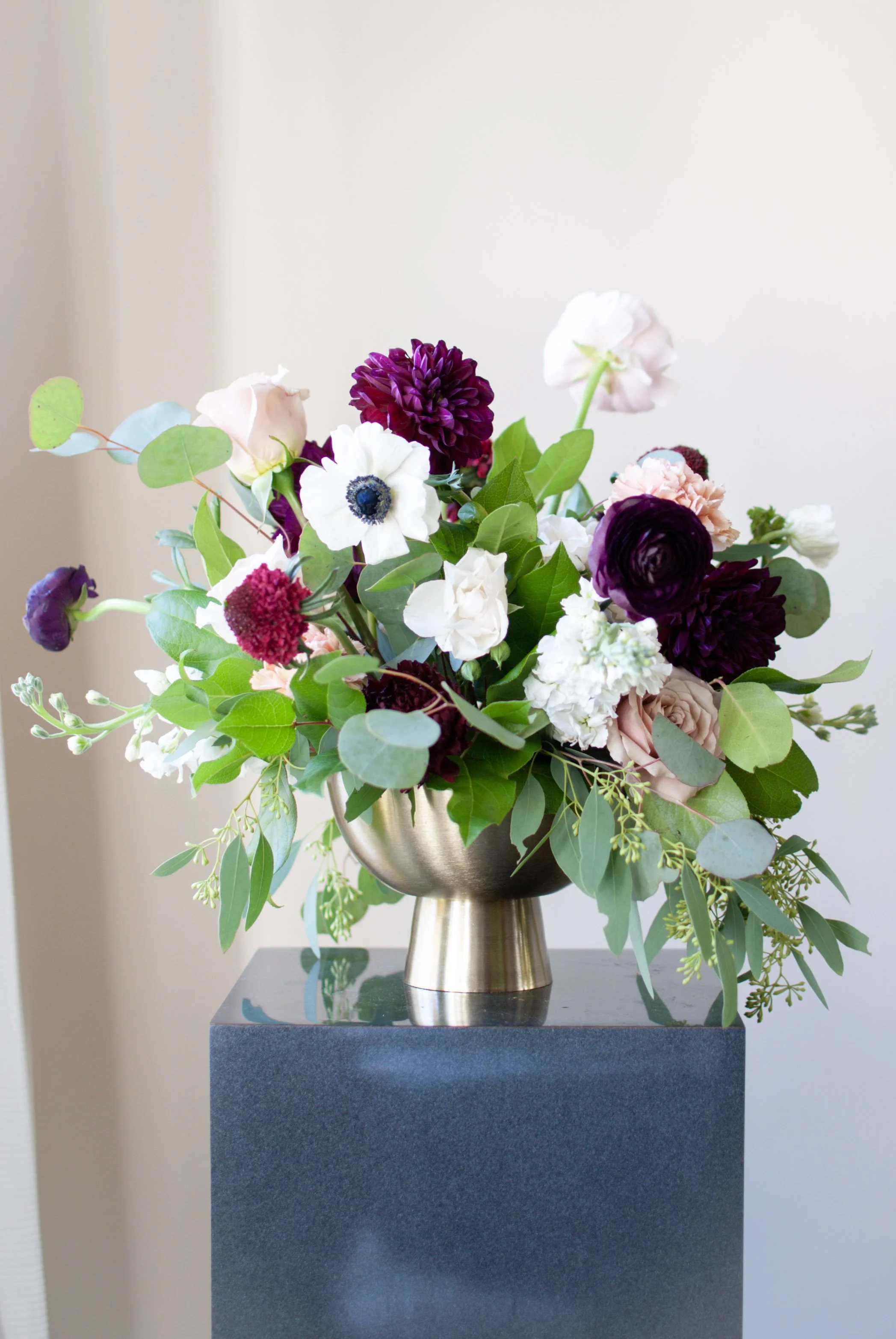 A bouquet of mixed flowers with purple, white, and blush pink blooms in a metallic gold vase on a dark pedestal against a plain light-colored wall.
