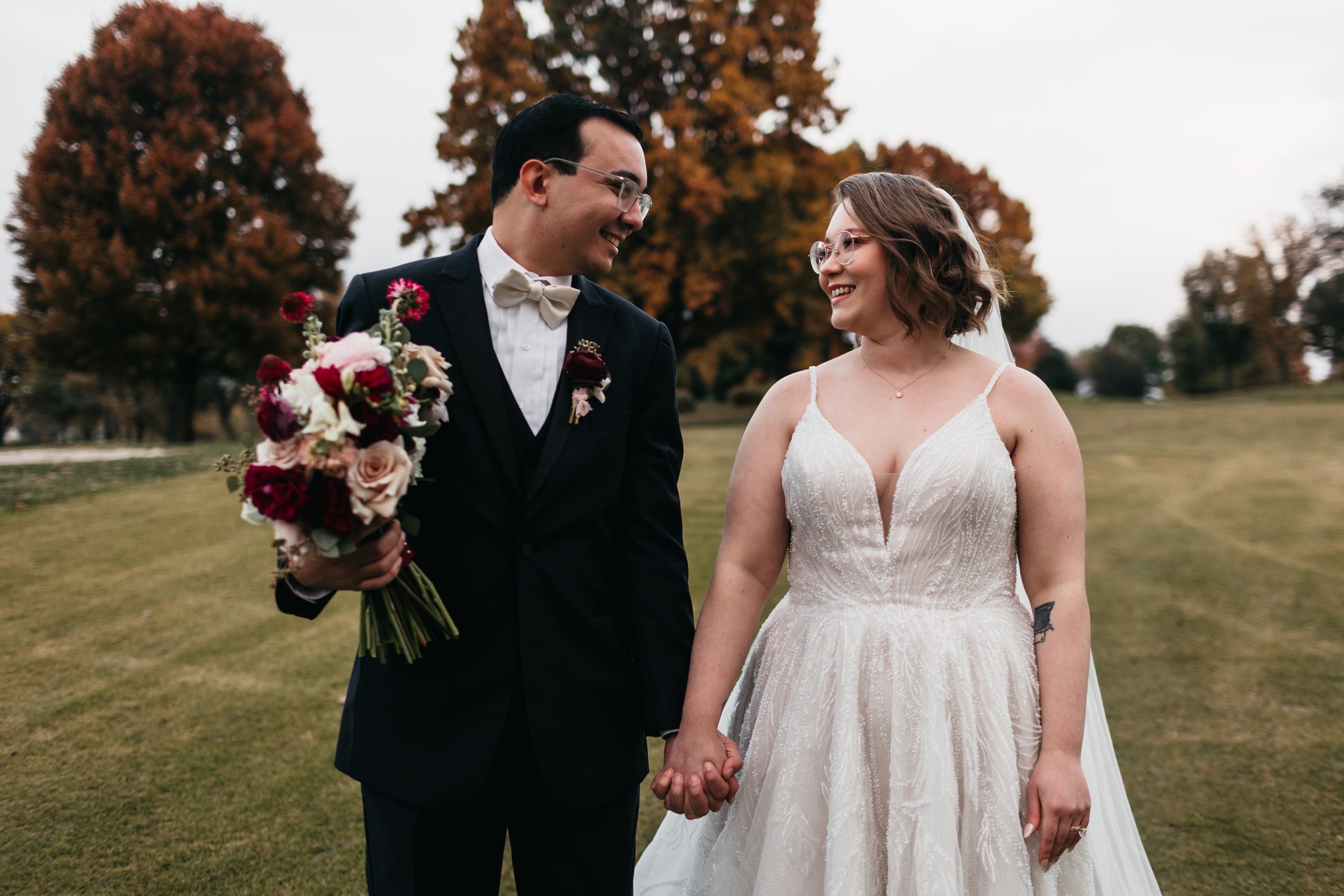 A newlywed couple holding hands and smiling at each other in a park with autumn trees in the background. Bouquet by St. Louis florist, Suncrest Blooms
