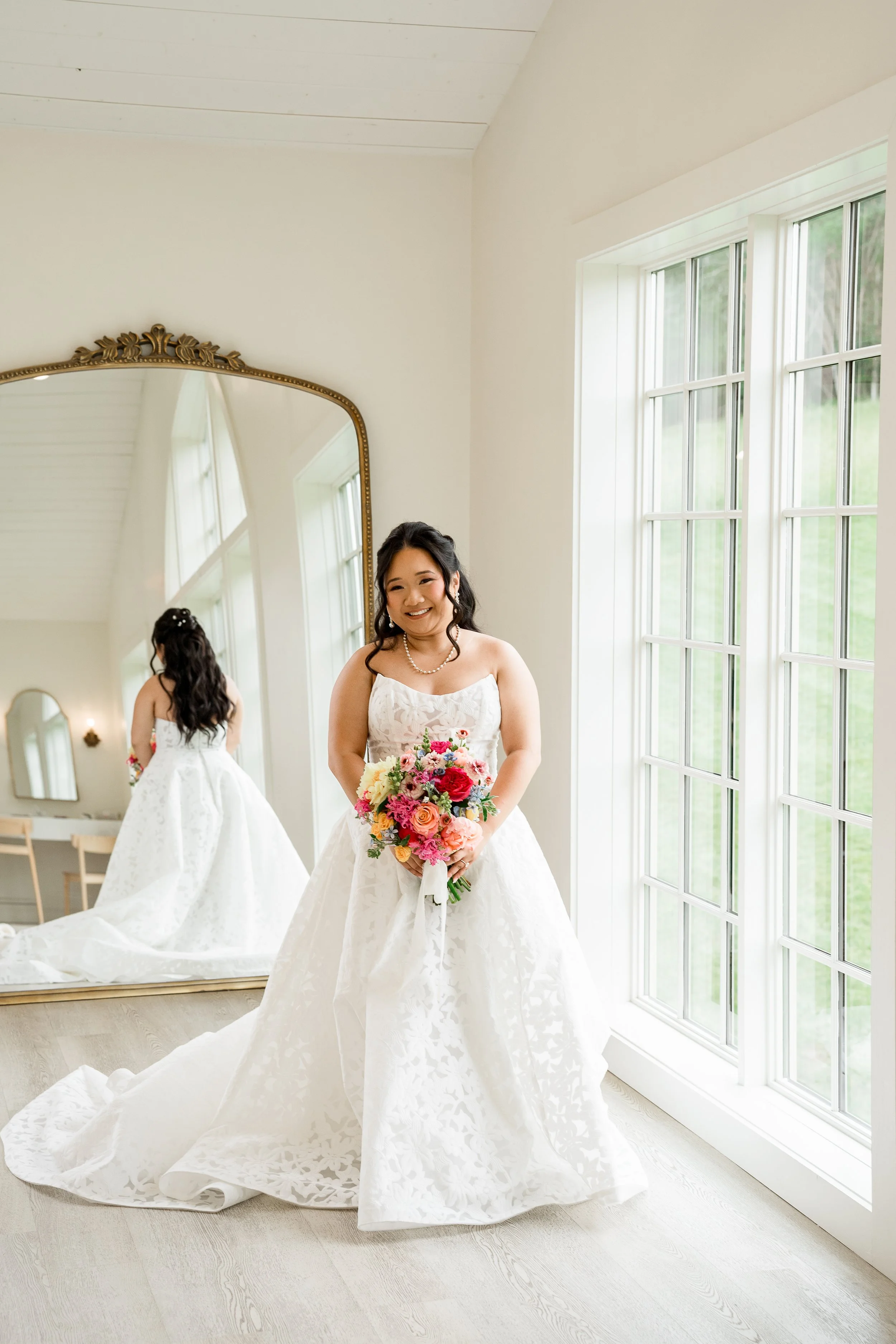 A bride stands in front of a large mirror smiling at the camera while holding her bridal bouquet. Florals by St. Louis Wedding Florist, Suncrest Blooms. Venue is Westwind Hills.