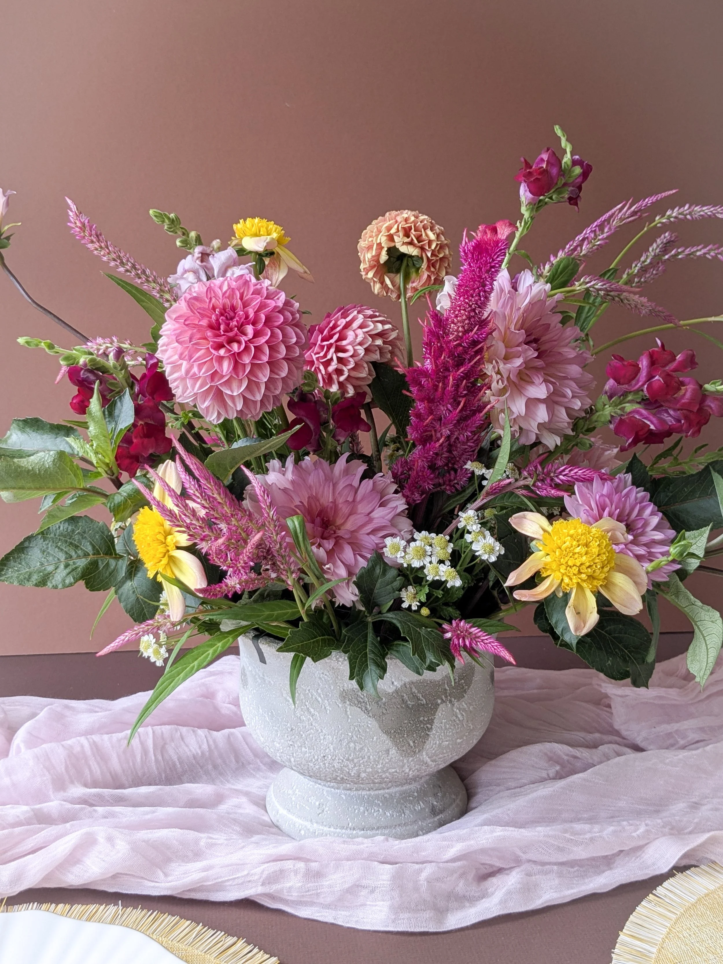 A vibrant bouquet of pink, yellow, and purple flowers in a textured white vase on a pink tablecloth, with a pink background.
