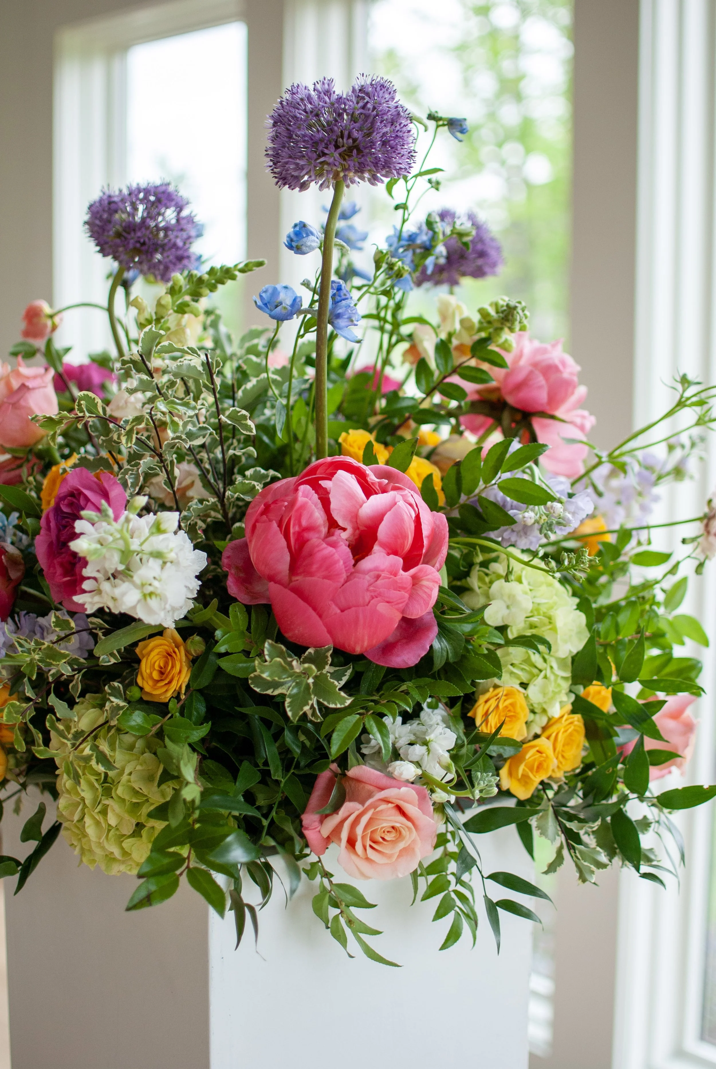 A vibrant bouquet of mixed flowers including pink roses, purple allium, blue gentians, yellow roses, white hydrangeas, and variegated greenery, arranged indoors near a window. Designed by St. Louis florist, Suncrest Blooms.