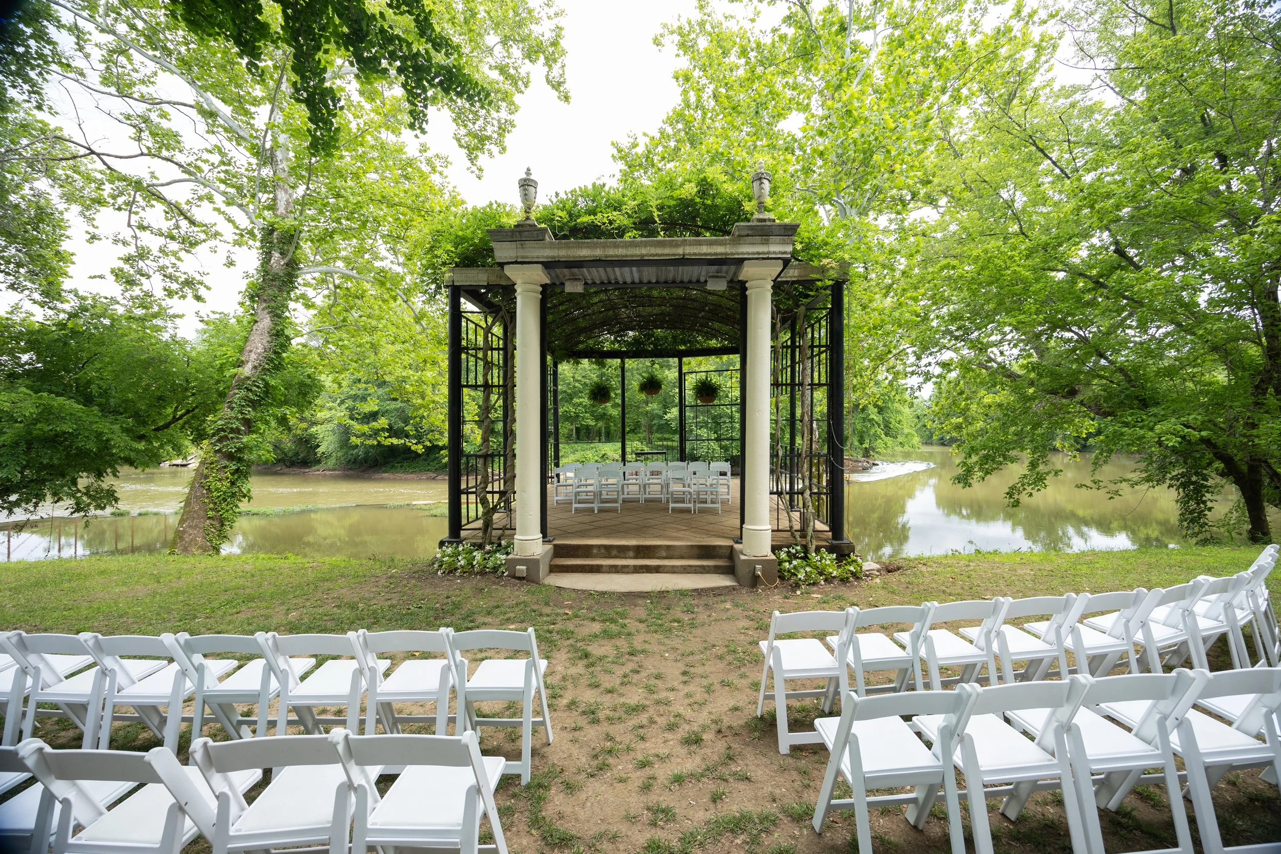 Outdoor wedding ceremony at Lalumondiere in St. Louis, Missouri, setup by the river with white chairs arranged facing a decorated gazebo with columns and hanging flower baskets, surrounded by green trees and calm water. St. Louis Florist.