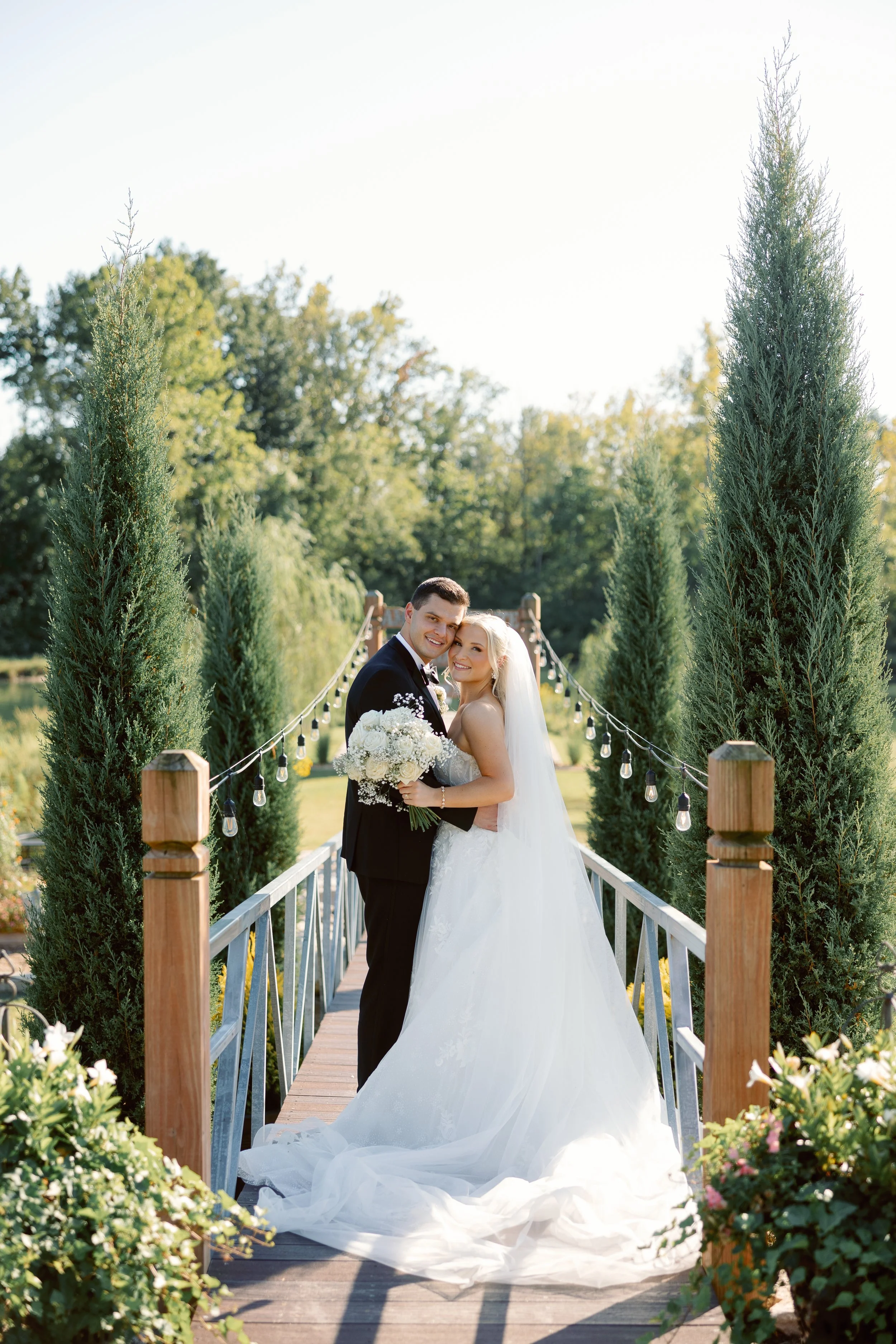 A newlywed couple smiling and embracing on a wooden bridge with string lights, surrounded by tall green trees and flowering bushes, during daytime outdoor wedding. Florals by St. Louis Florist, Suncrest Blooms.