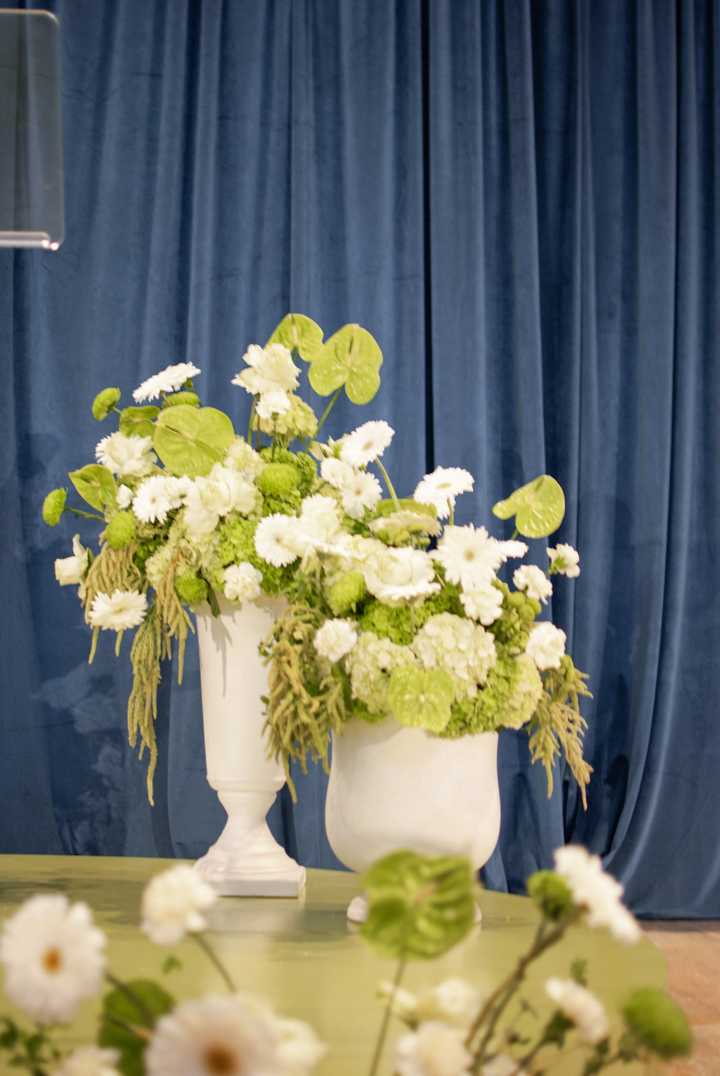 A large floral arrangement with white and light green flowers and green leaves, placed in two white vases. The background features blue curtains, and the arrangement is set on a green surface. St. Louis Florist. St. Louis Art Museum events.