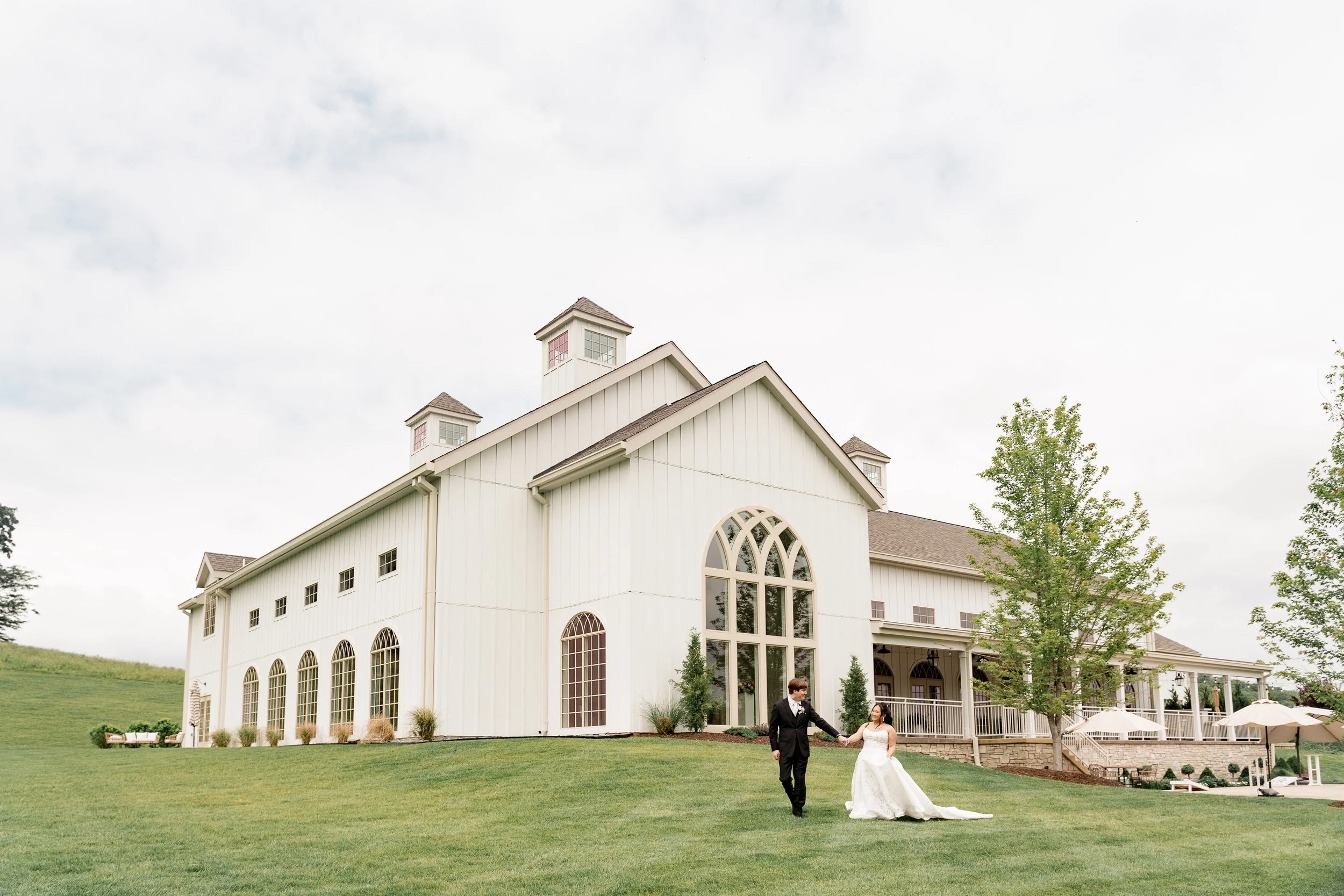 A bride and groom walk on a grassy field in front of Westwind Hills, a luxury wedding venue in St. Louis, Missouri.