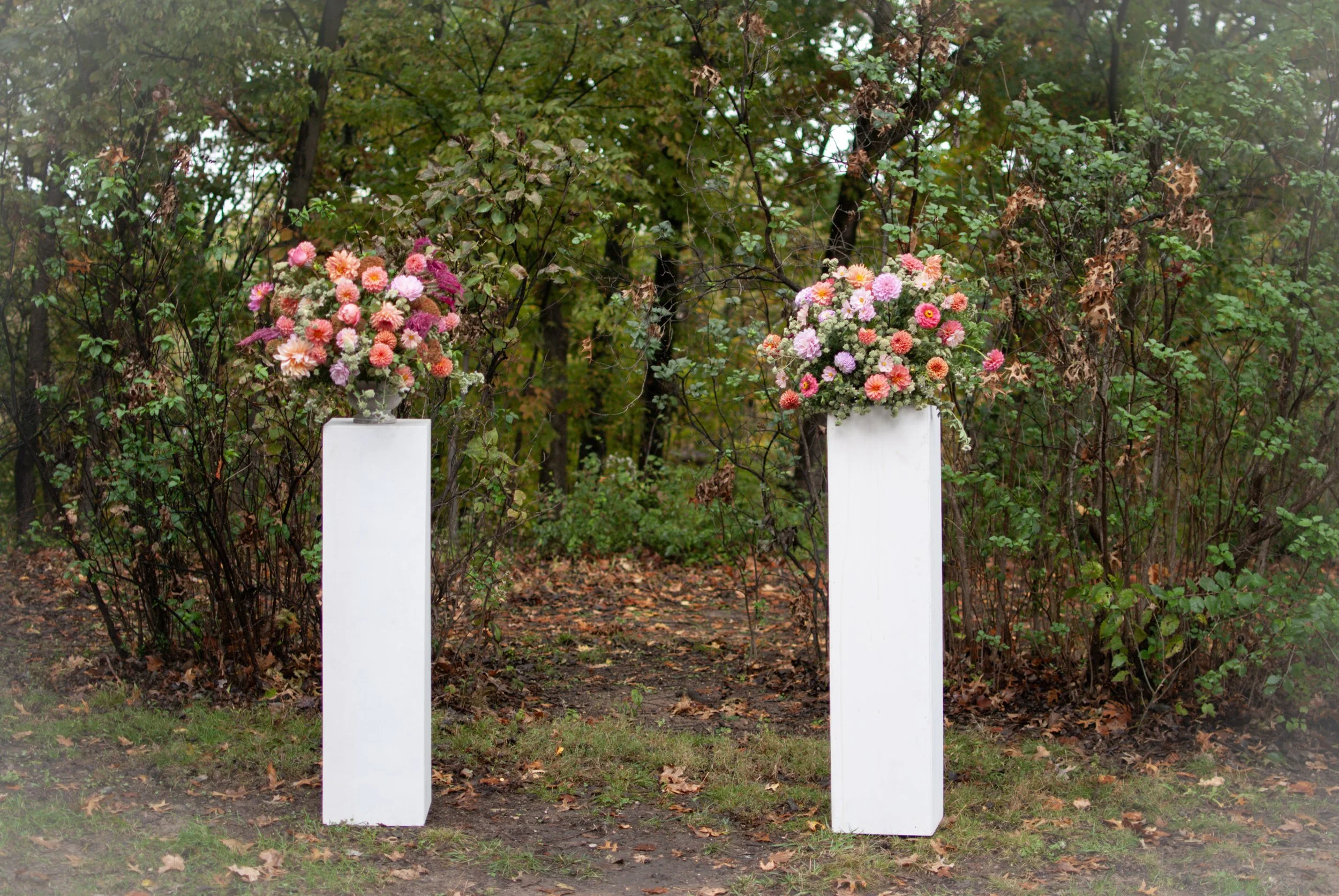 Two white pedestals with large bouquets of pink, peach, and white flowers are positioned outdoors against a backdrop of trees and foliage.