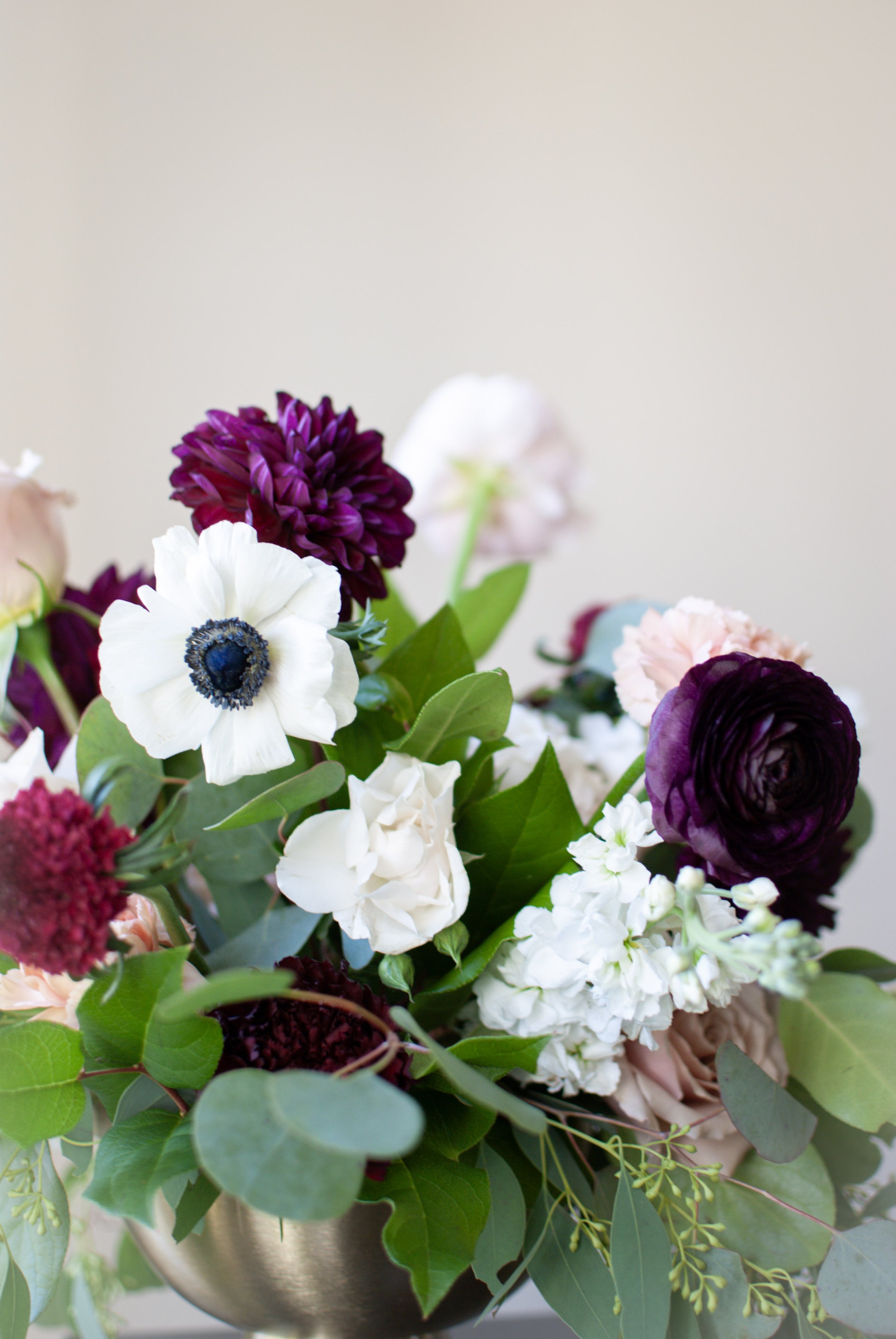 A colorful bouquet of flowers including white anemones, dark purple ranunculus, burgundy flowers, light pink roses, and eucalyptus leaves in a metallic vase.