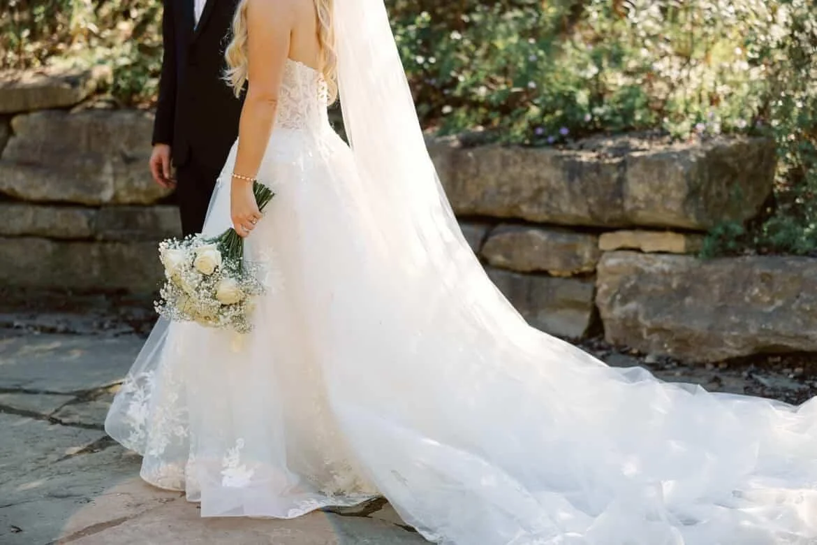 Bride in a white wedding gown holding a bouquet of white flowers, with a man in a black suit partly visible, outdoors surrounded by greenery and rocks.