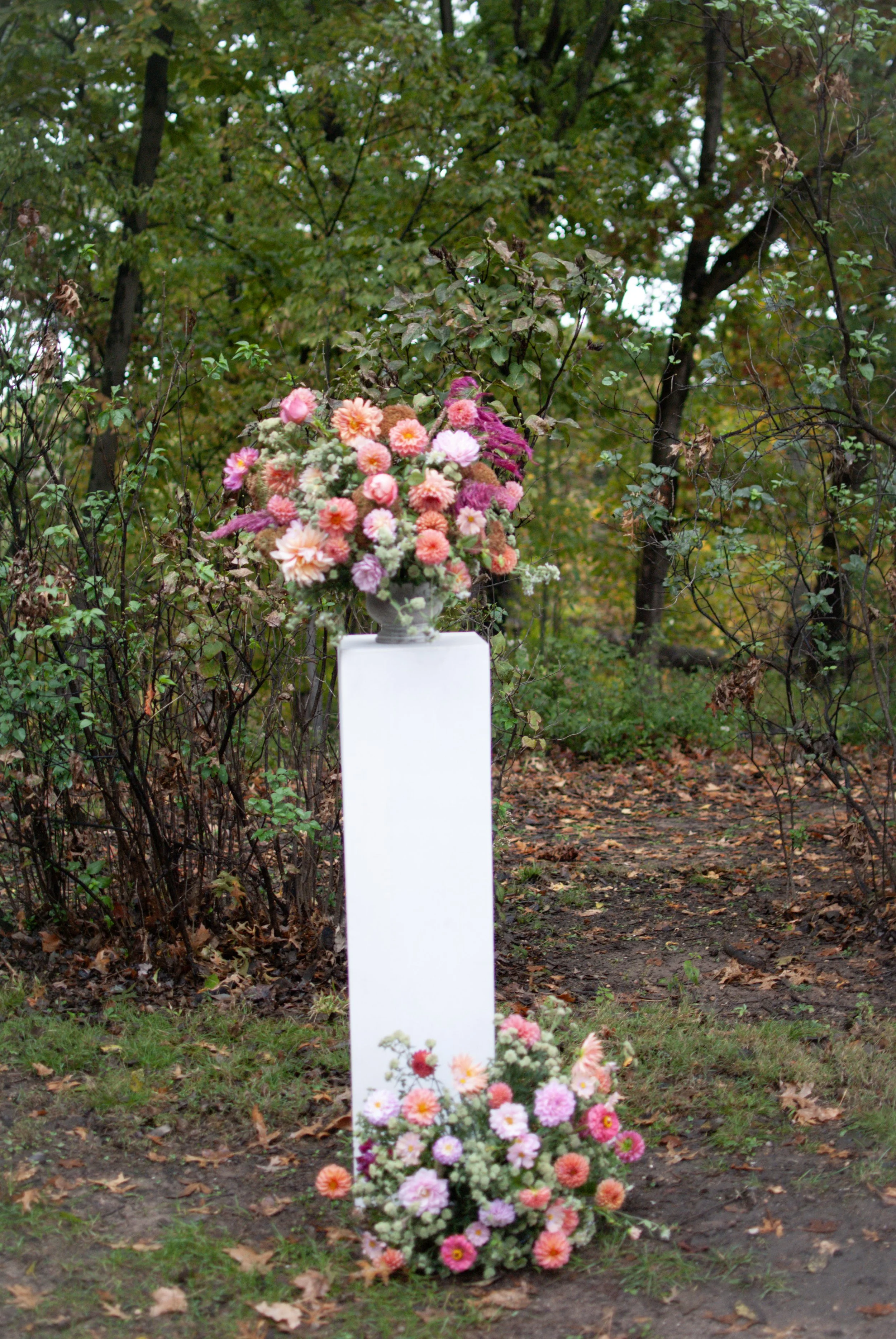 A large floral arrangement with pink, white, and orange flowers on a white pedestal, with smaller flower arrangements at its base, set outdoors among trees and fallen leaves.