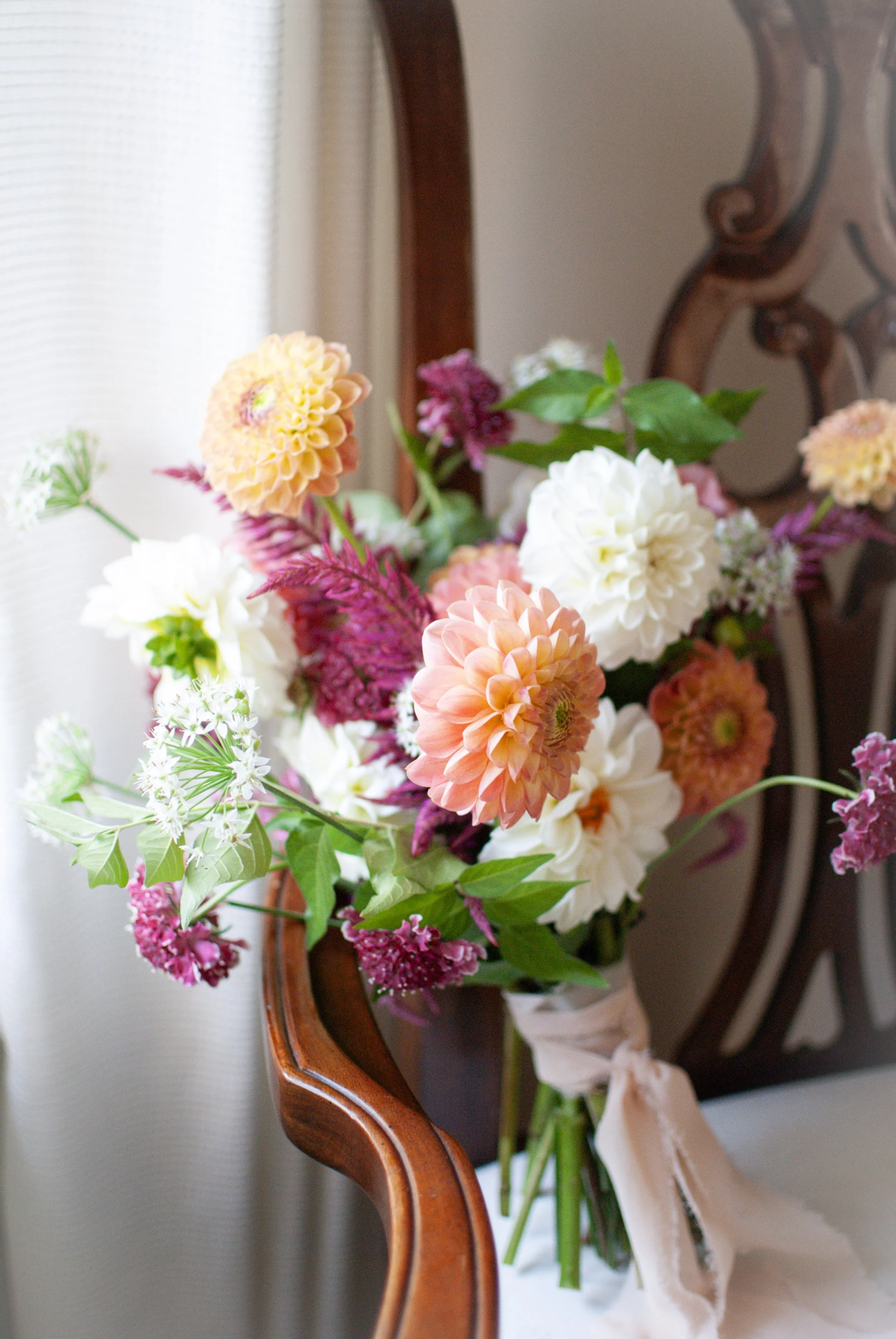 A bouquet of colorful flowers resting on a wooden chair with white fabric and soft curtains in the background.