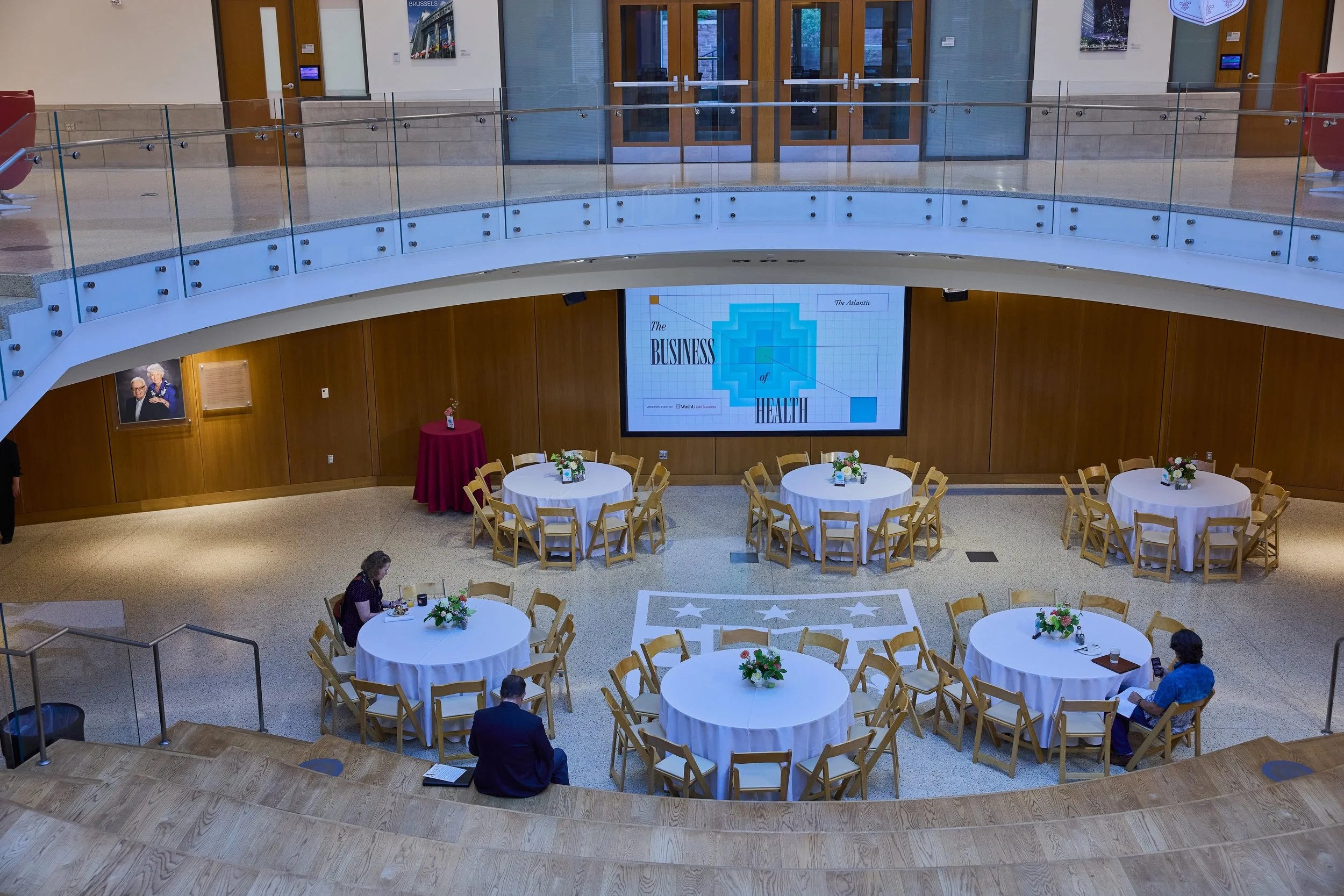 Conference room setup with round tables covered in white tablecloths and floral centerpieces, a large screen displaying 'The Business of Health,' and a few people seated. The room has wooden walls, a balcony with glass railing, and a polished floor with stars and stripes design.