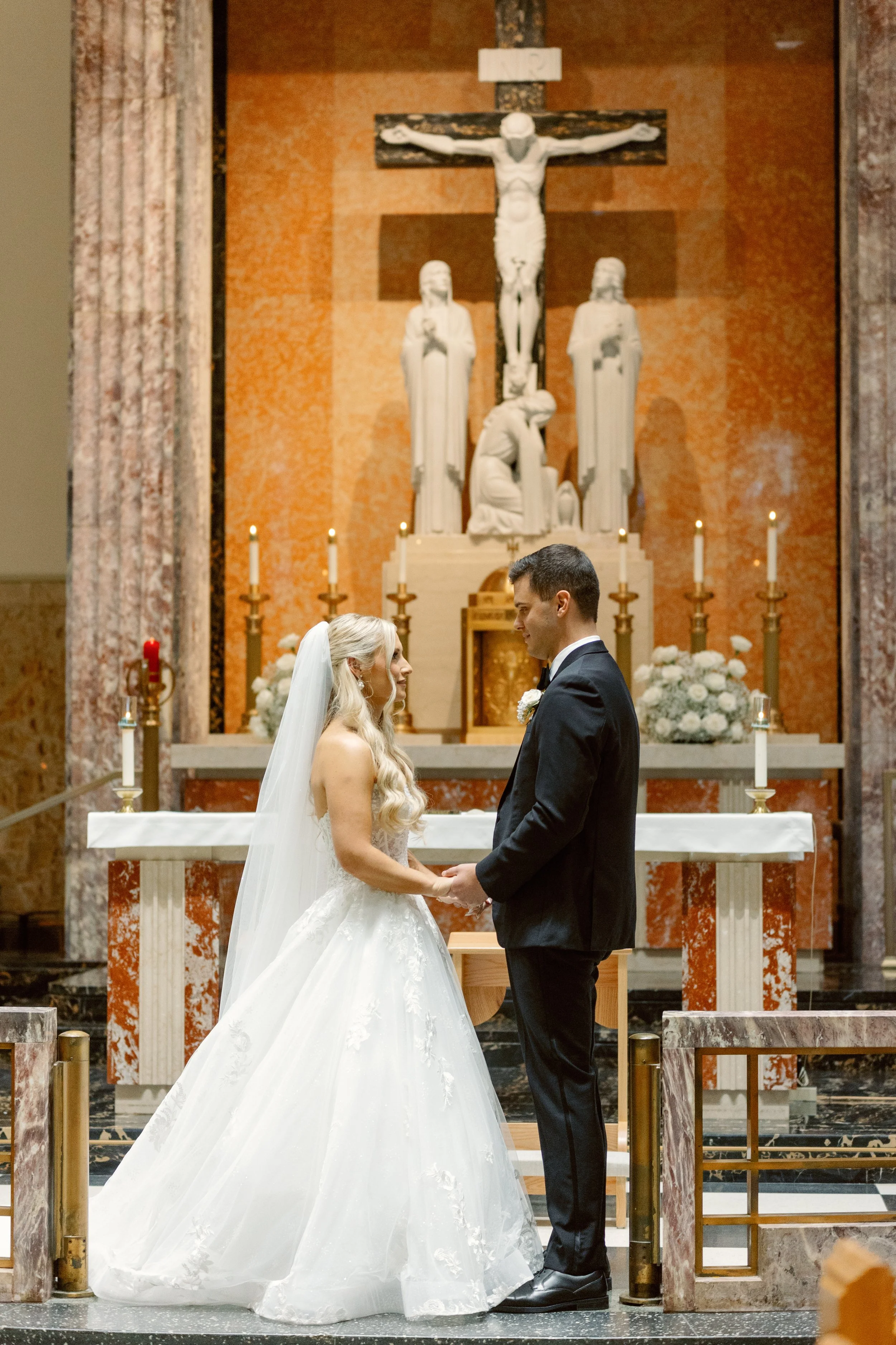 A bride and groom standing face-to-face holding hands during their wedding ceremony inside a church with religious statues and candles in the background.