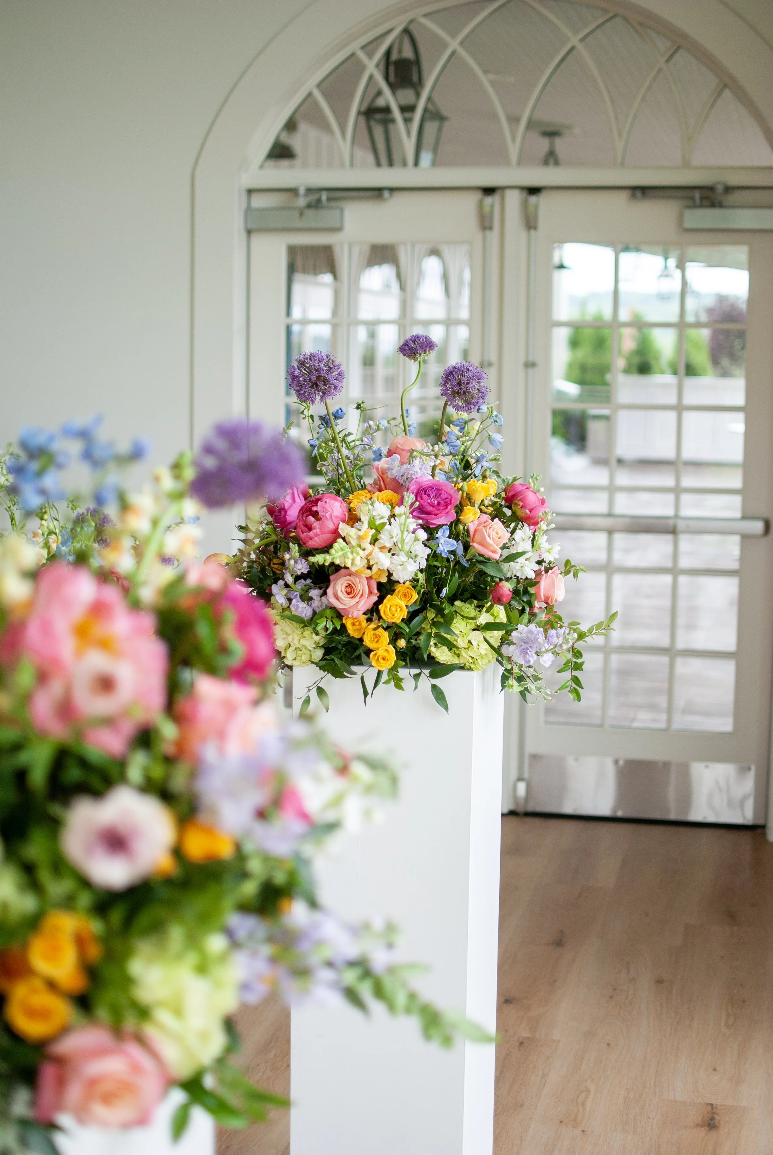 Vases of colorful flowers in front of a glass door with white framing, leading to an outdoor porch with a railing and trees visible through the windows. Designed by garden-style St. Louis florist, Suncrest Blooms.