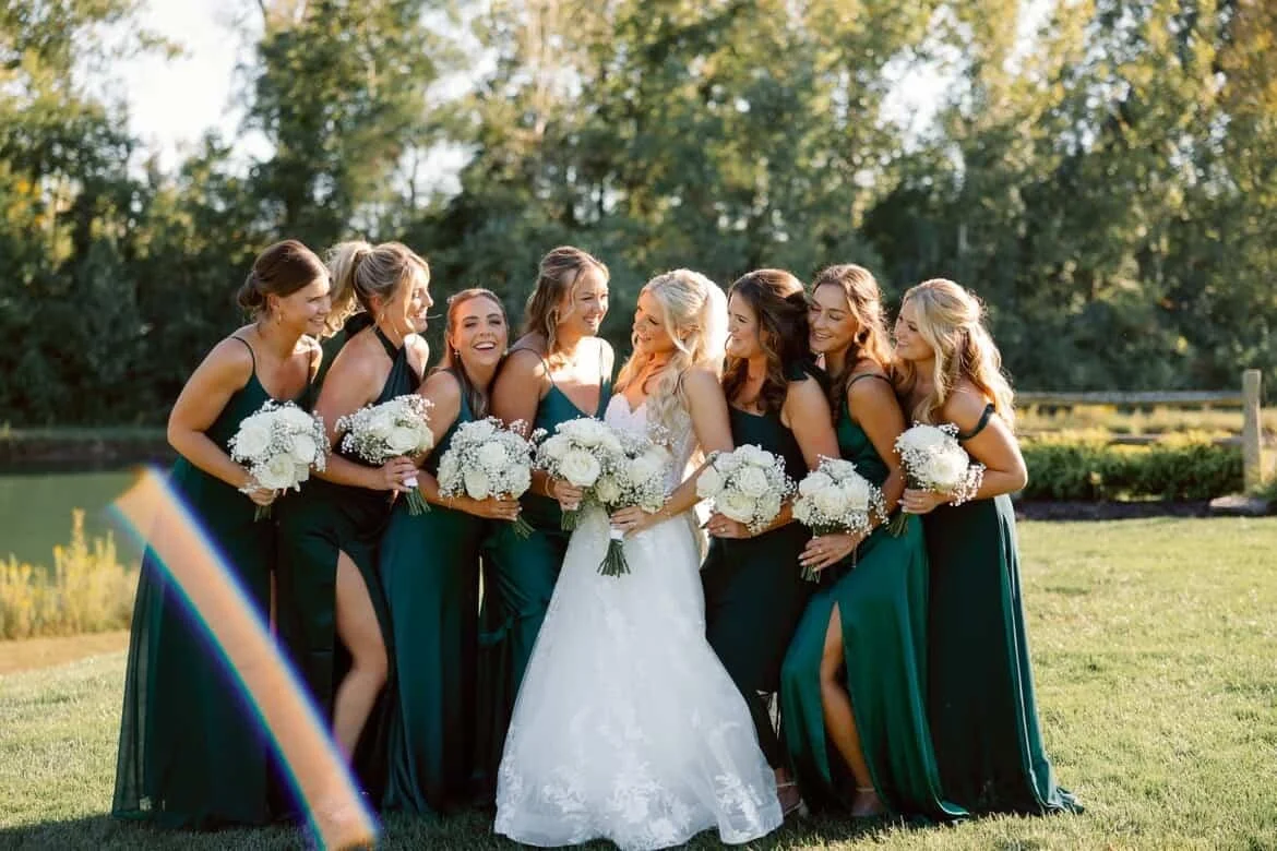 A bride and seven bridesmaids standing outdoors on a grassy area with trees in the background. They are dressed in matching dark green dresses and holding white bouquets and smiling. Florals designed by St. Louis Florist, Suncrest Blooms.