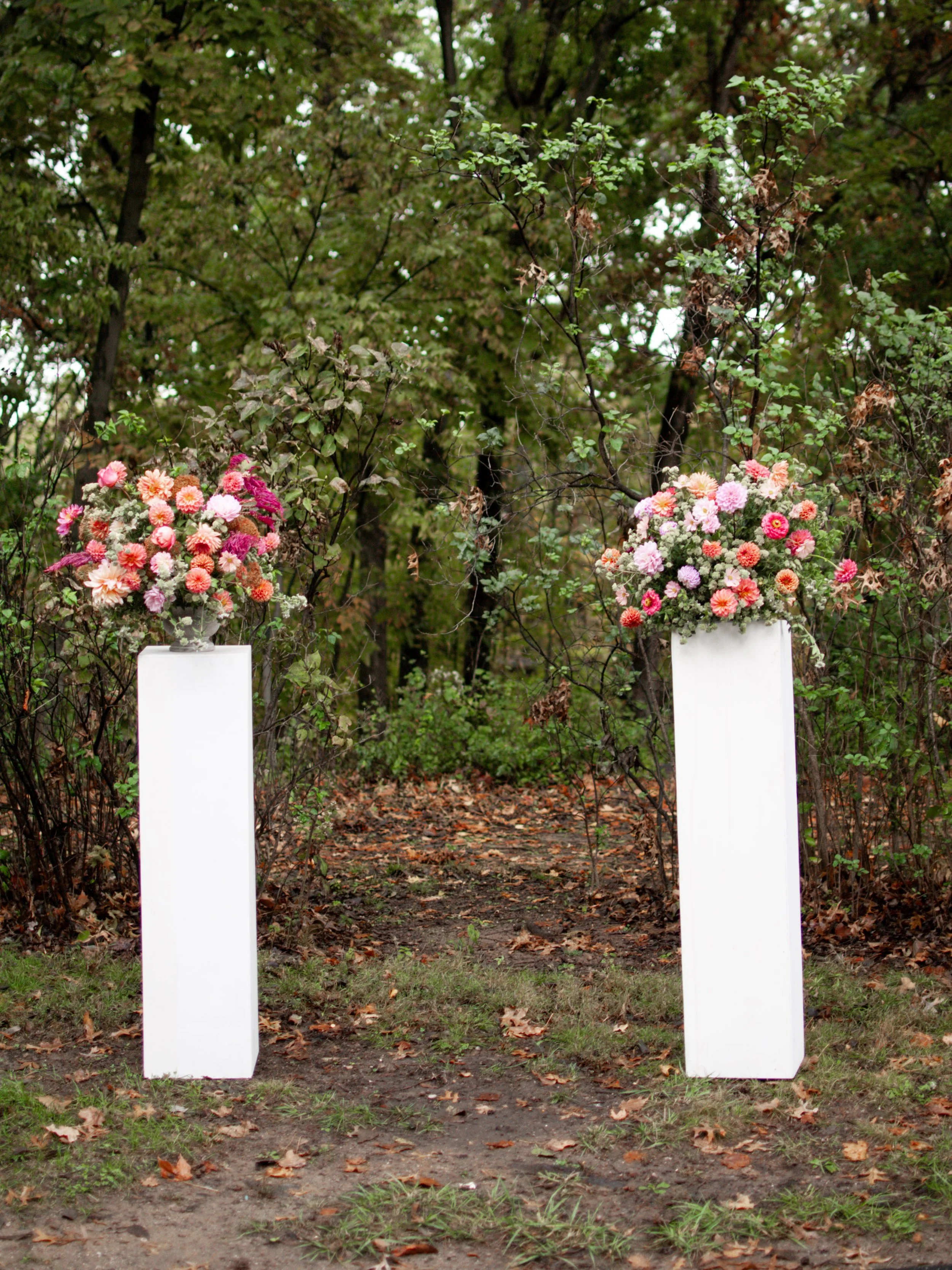 Two white pedestals with large flower arrangements of pink and red flowers placed outdoors in front of trees and foliage. Florals by St. Louis Florist, Suncrest Blooms.