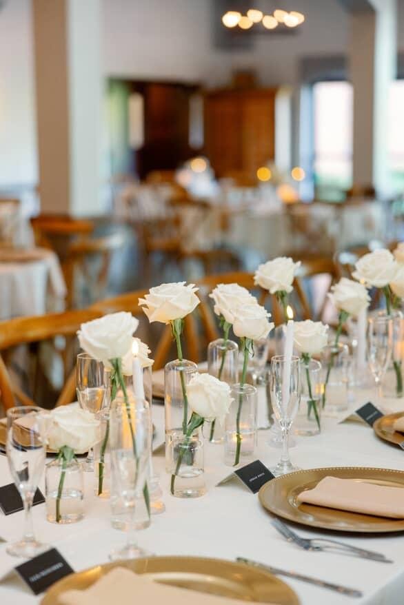 A beautifully set wedding or event reception table with white roses in tall glass vases, champagne glasses, gold plates, and beige napkins, in a well-lit venue with wooden chairs and tables.