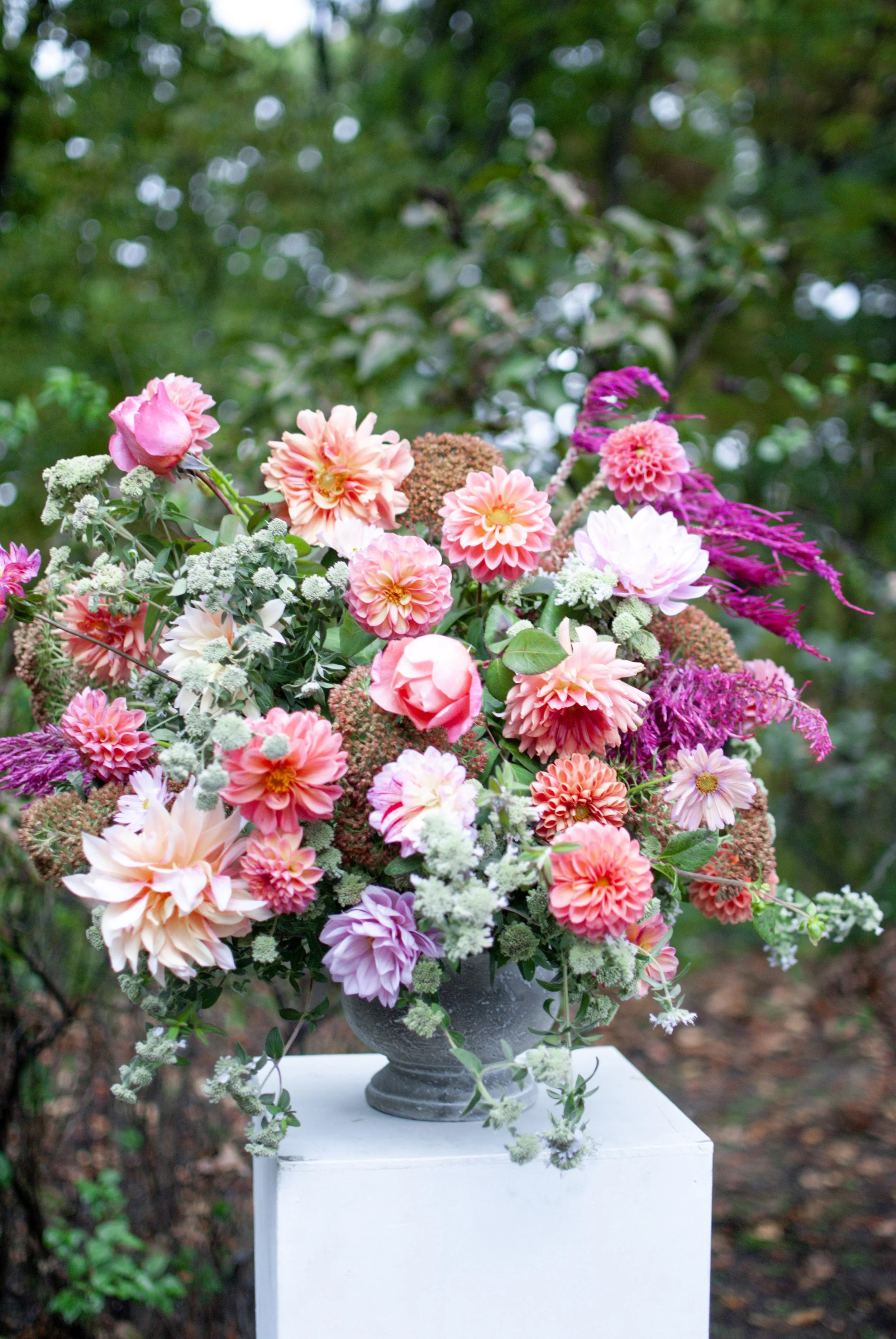 Colorful bouquet of pink, peach, and purple flowers arranged in a gray ceramic vase, placed on a white pedestal outdoors with a blurred green foliage background.