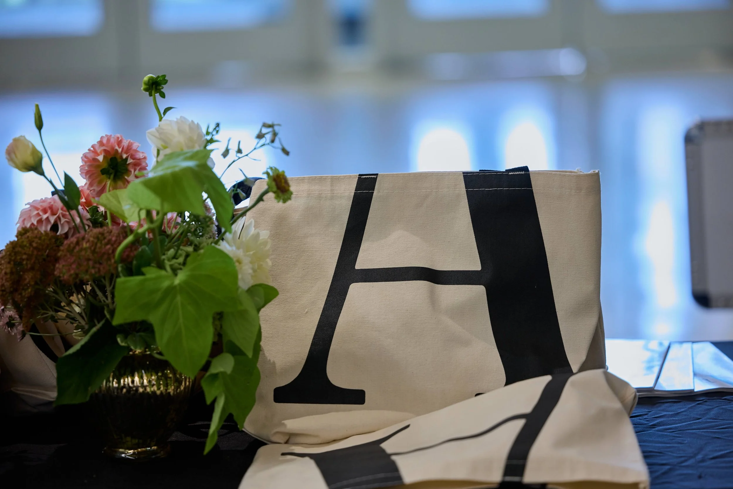 A tote bag with a large black letter 'A' on it, a bouquet of mixed flowers, and a folded cloth with black lettering on a dark surface.