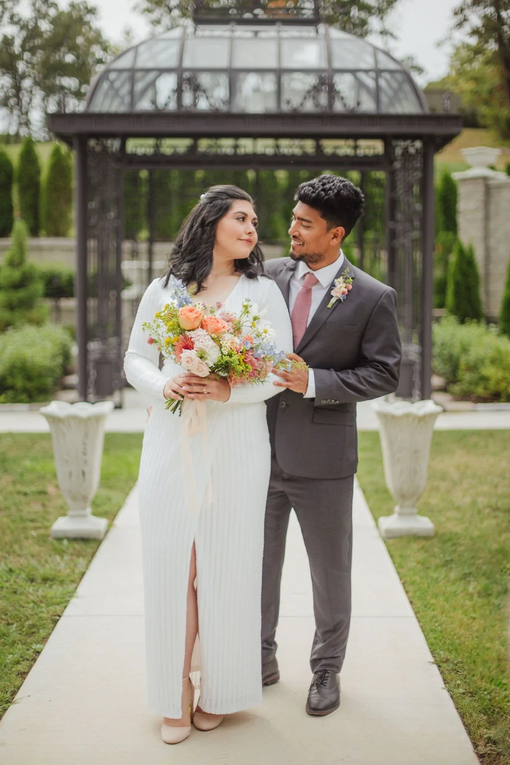 A bride and groom standing together outdoors, with the bride holding a bouquet of flowers, in a garden setting with a greenhouse structure in the background. Florals designed by St. Louis Florist, Suncrest Blooms.