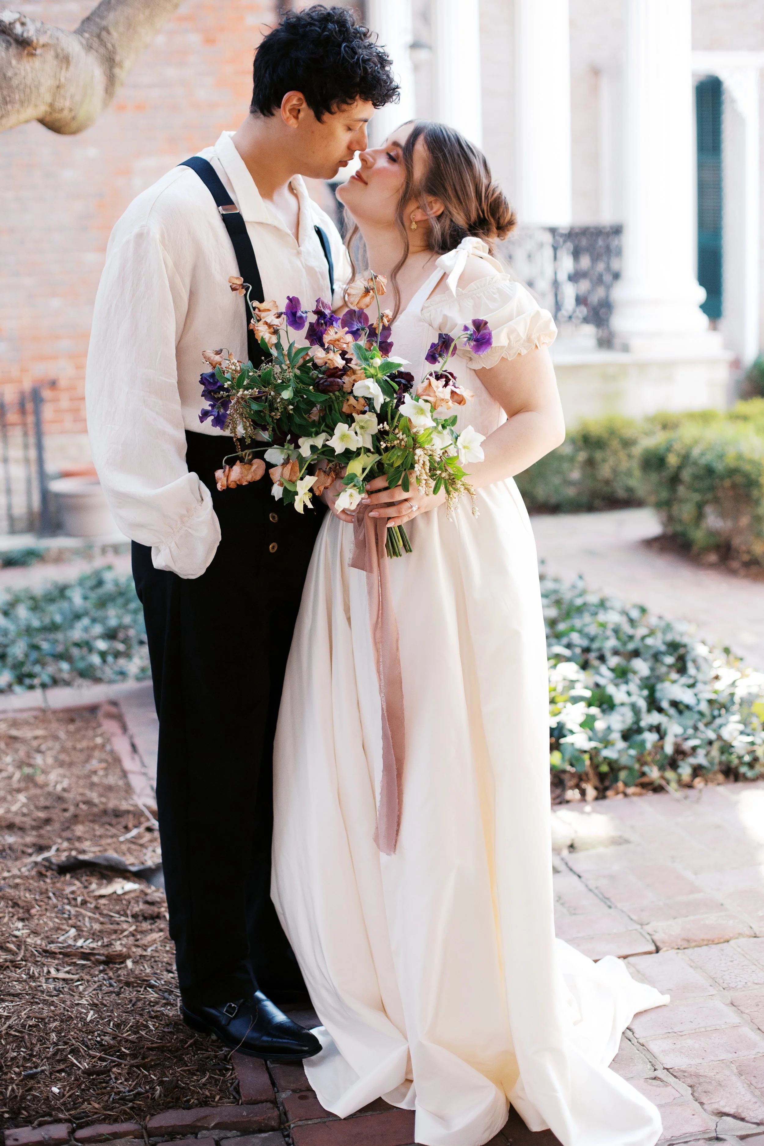 A couple dressed in vintage fashion standing close on a sidewalk, holding a bouquet of colorful flowers, gazing at each other with their noses touching. Designed by St. Louis wedding and event florist, Suncrest Blooms.