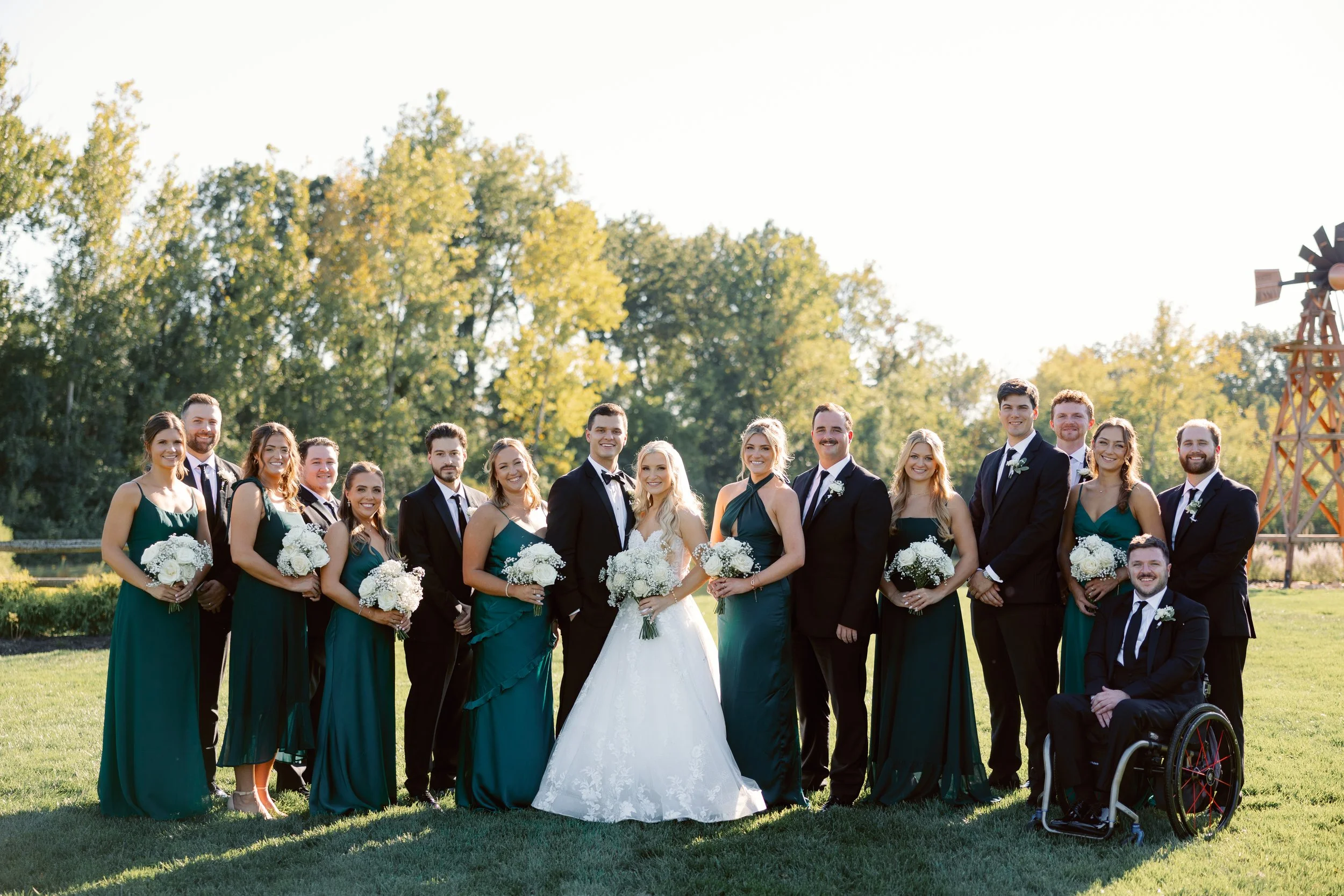 Wedding party outdoors, including bride, groom, bridesmaids, groomsmen, and a person in a wheelchair, standing on grass with trees and a windmill in the background.