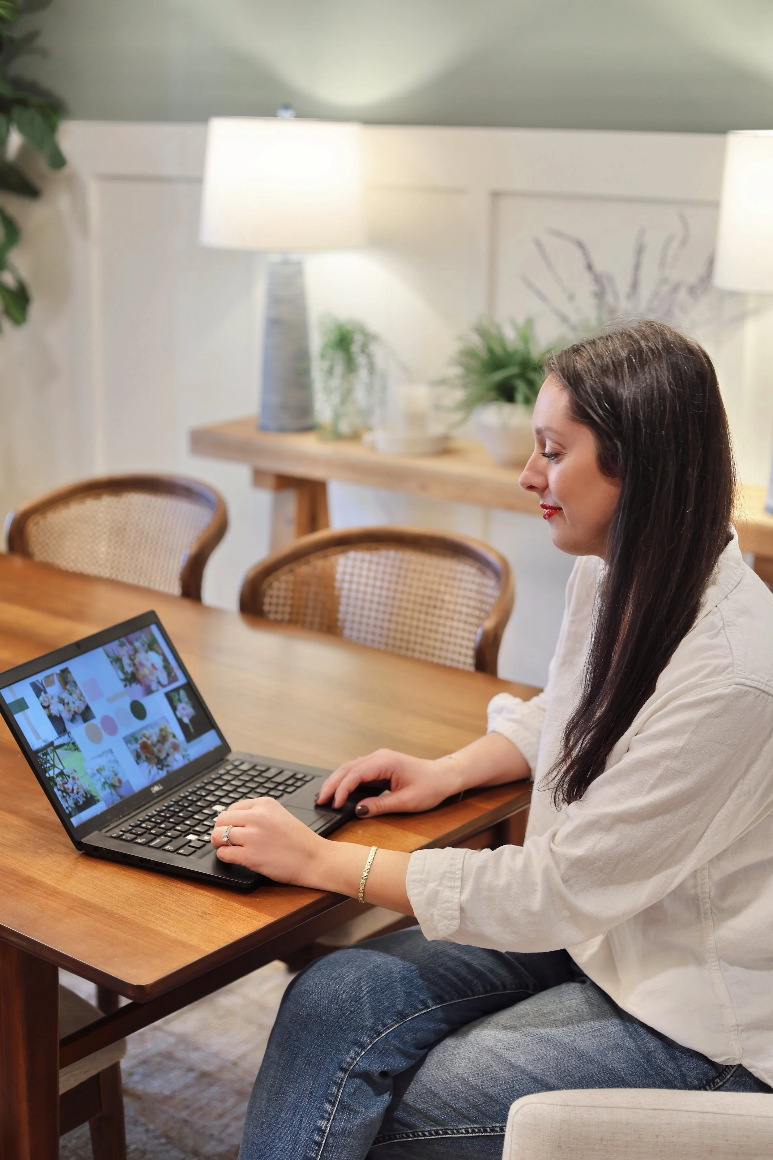 Woman sitting at a wooden dining table using a laptop, in a cozy, well-lit room with lamps and plants. This is Dawn Gross, of Suncrest Blooms. Suncrest Blooms is a wedding and event florist based in St. Louis, Missouri.
