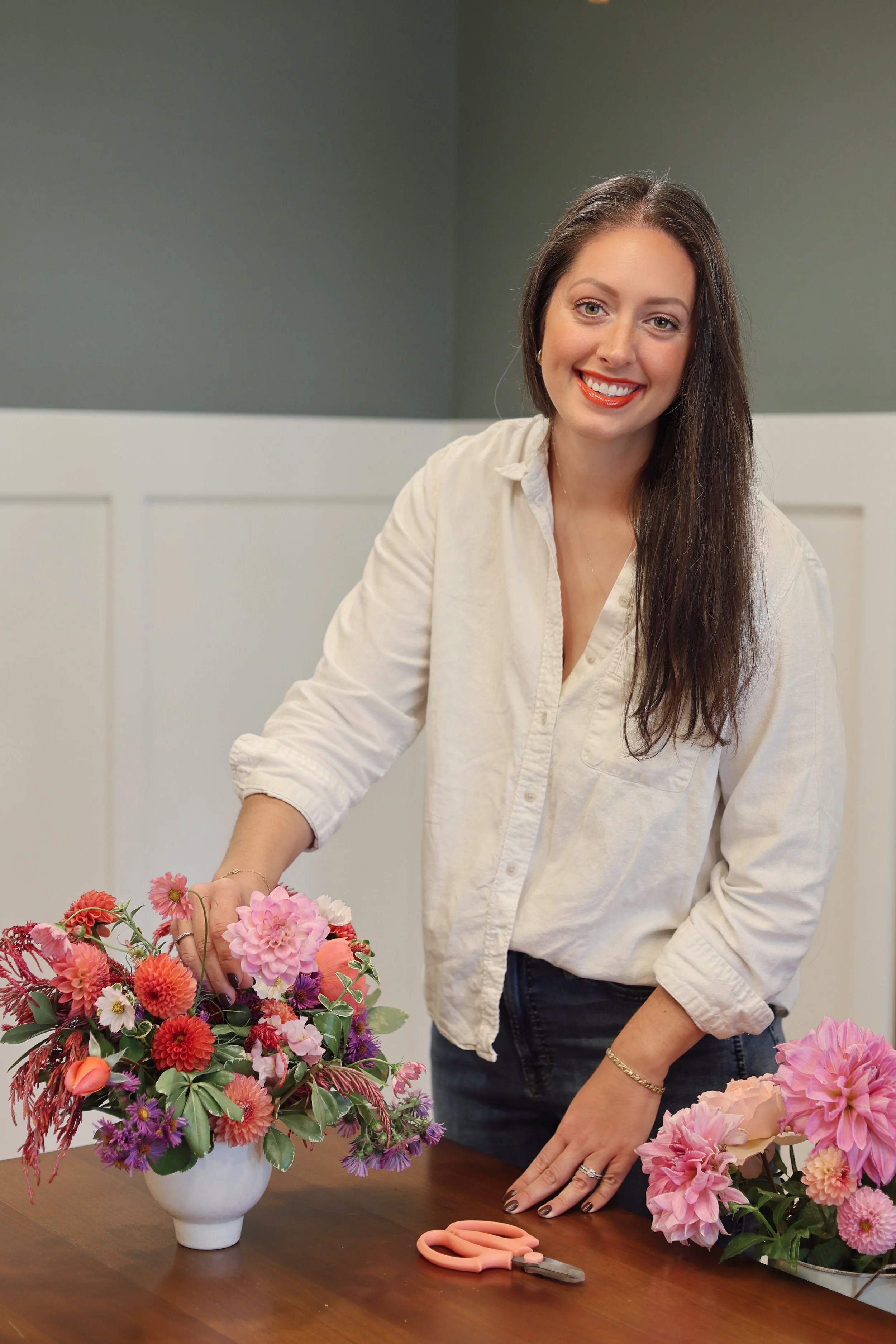 A woman with long brown hair and a white shirt arranging pink and red flowers in a white vase on a wooden table, with a pair of pink scissors nearby. This is Dawn Gross of Suncrest Blooms, St. Louis Florist for weddings & events.