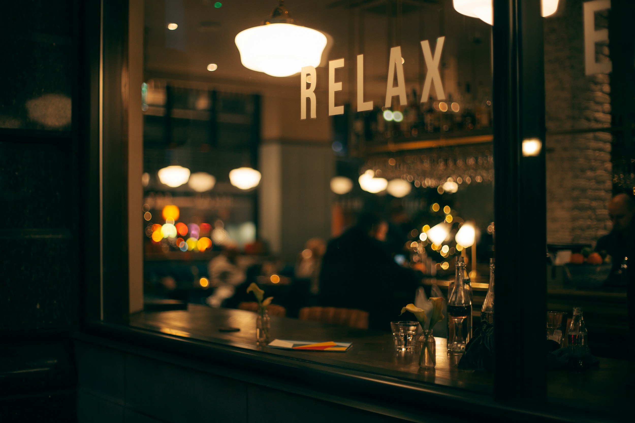 A cozy restaurant scene viewed through a window with the word 'RELAX' on it. Inside, dim lighting and hanging lamps create a warm, inviting atmosphere with people dining and drinking.