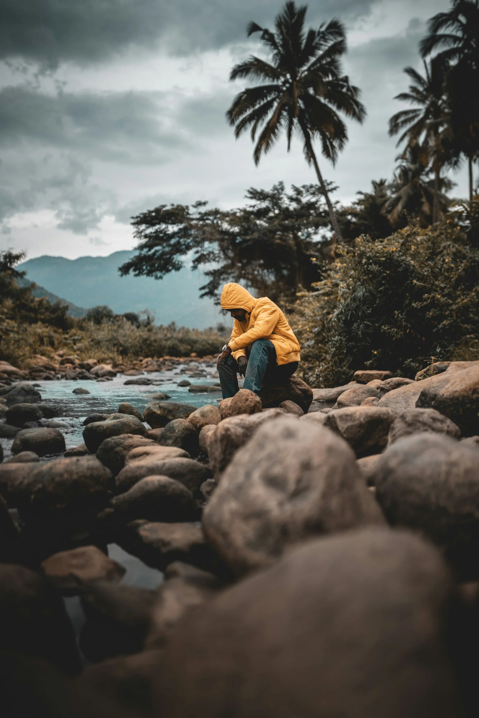 Person in a yellow jacket sitting on rocks by a river, with tropical trees and mountains under a cloudy sky in the background.