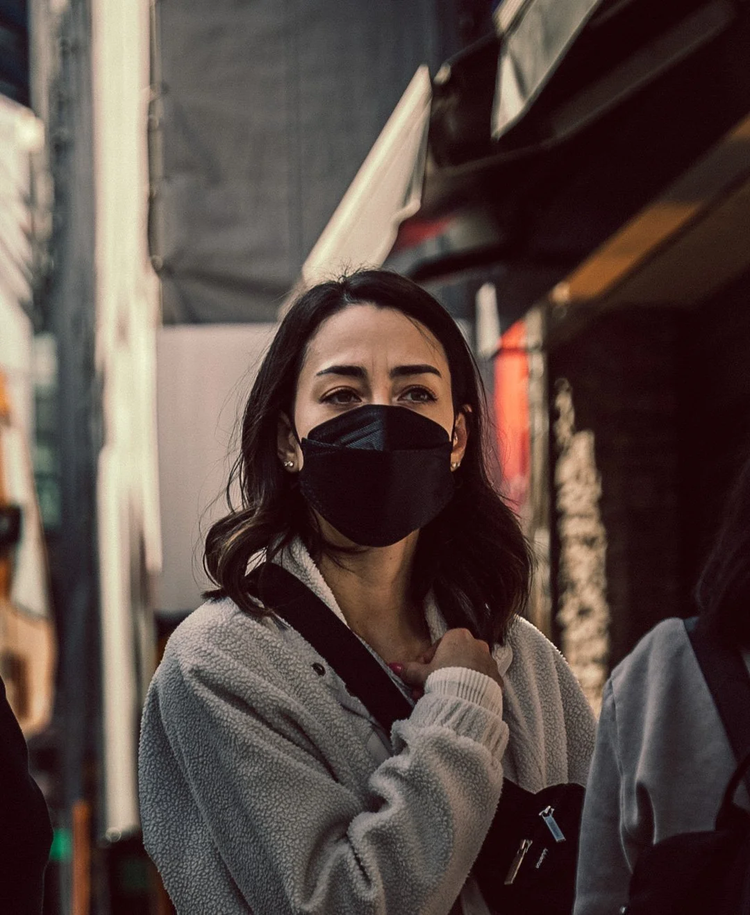 A woman with dark hair wearing a black face mask and a light-colored jacket in an urban setting at dusk.