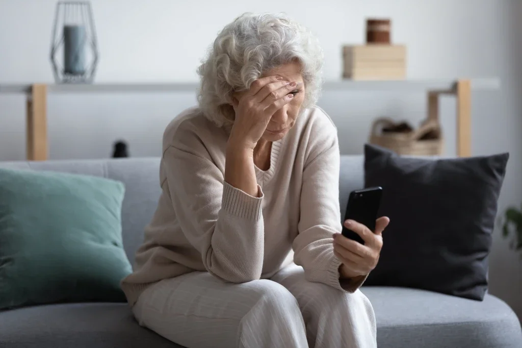 An elderly woman sitting on a couch, holding her forehead with one hand, looking at a smartphone in her other hand, in a living room with pillows and decor in the background.