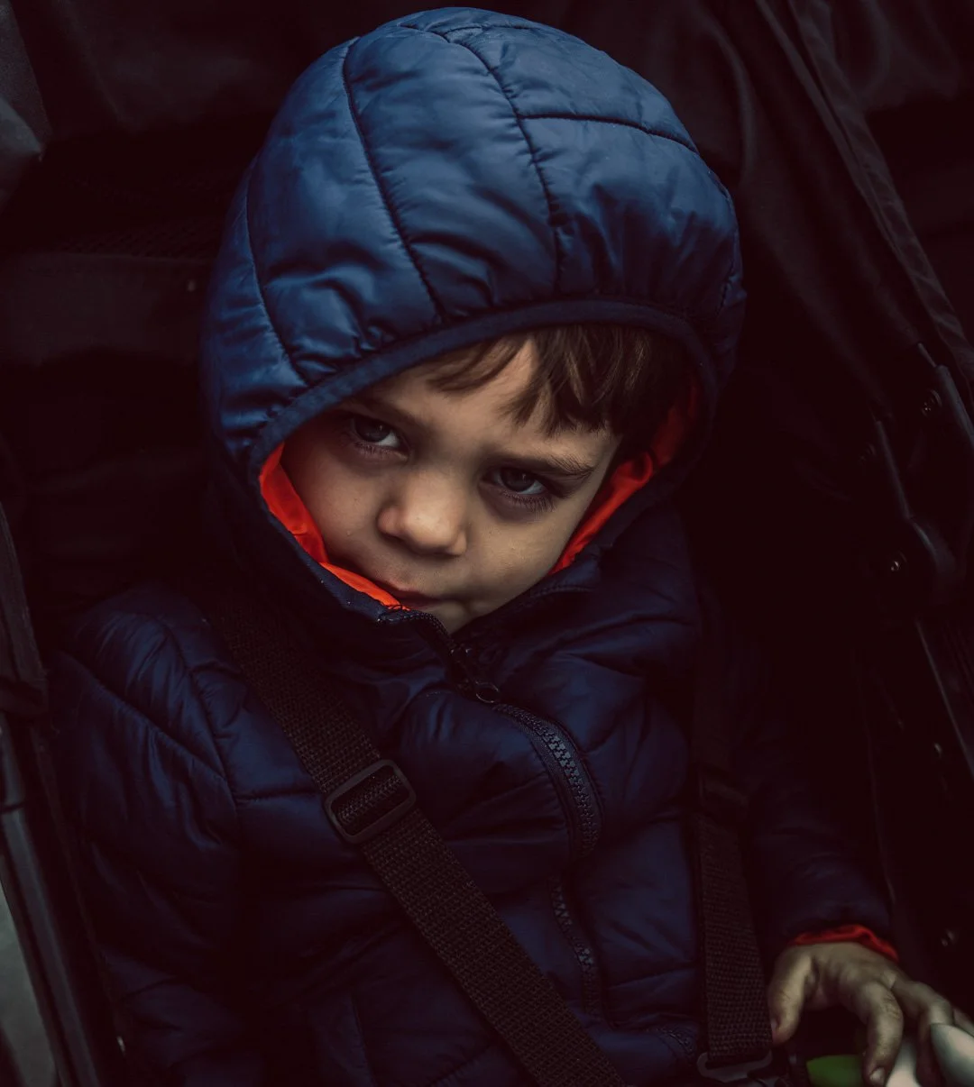 A young boy with light brown hair and blue eyes sitting in a stroller, wearing a navy blue winter jacket with an orange lining and a matching hood. He looks directly at the camera with a serious expression.