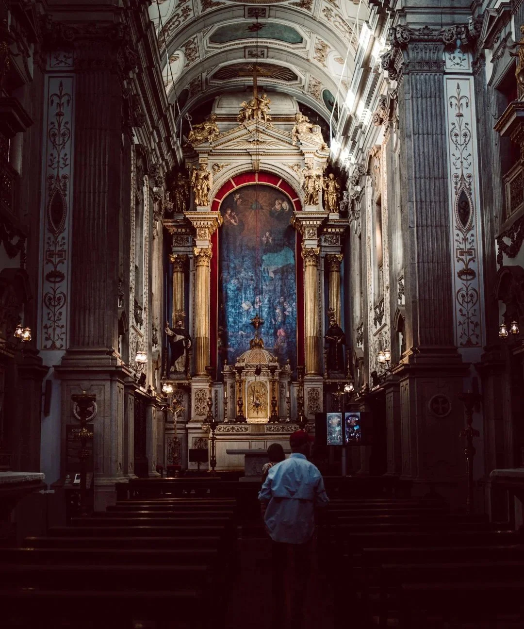 Interior of a grand, ornately decorated church with gold accents, columns, and religious artwork, including a large painting behind the altar and a crucifix in front. Two people stand near the steps leading to the altar.