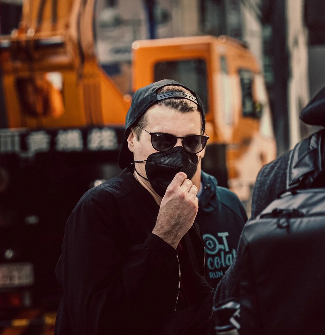 A man wearing black sunglasses, a black face mask, a black baseball cap worn backwards, and a black jacket, standing outdoors with construction vehicles in the background.
