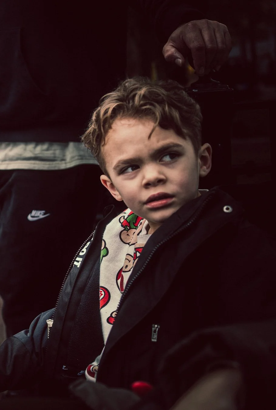 A young boy with curly hair, wearing a jacket and a graphic shirt featuring characters from Mario, looking to the side with a serious expression. An adult's hand is visible above him, holding a black object.
